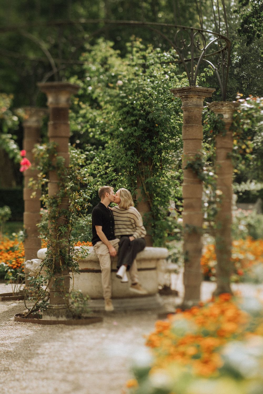 Couple sharing a romantic kiss in a lush garden pergola surrounded by colorful flowers and stone columns. Kasteel de Haar