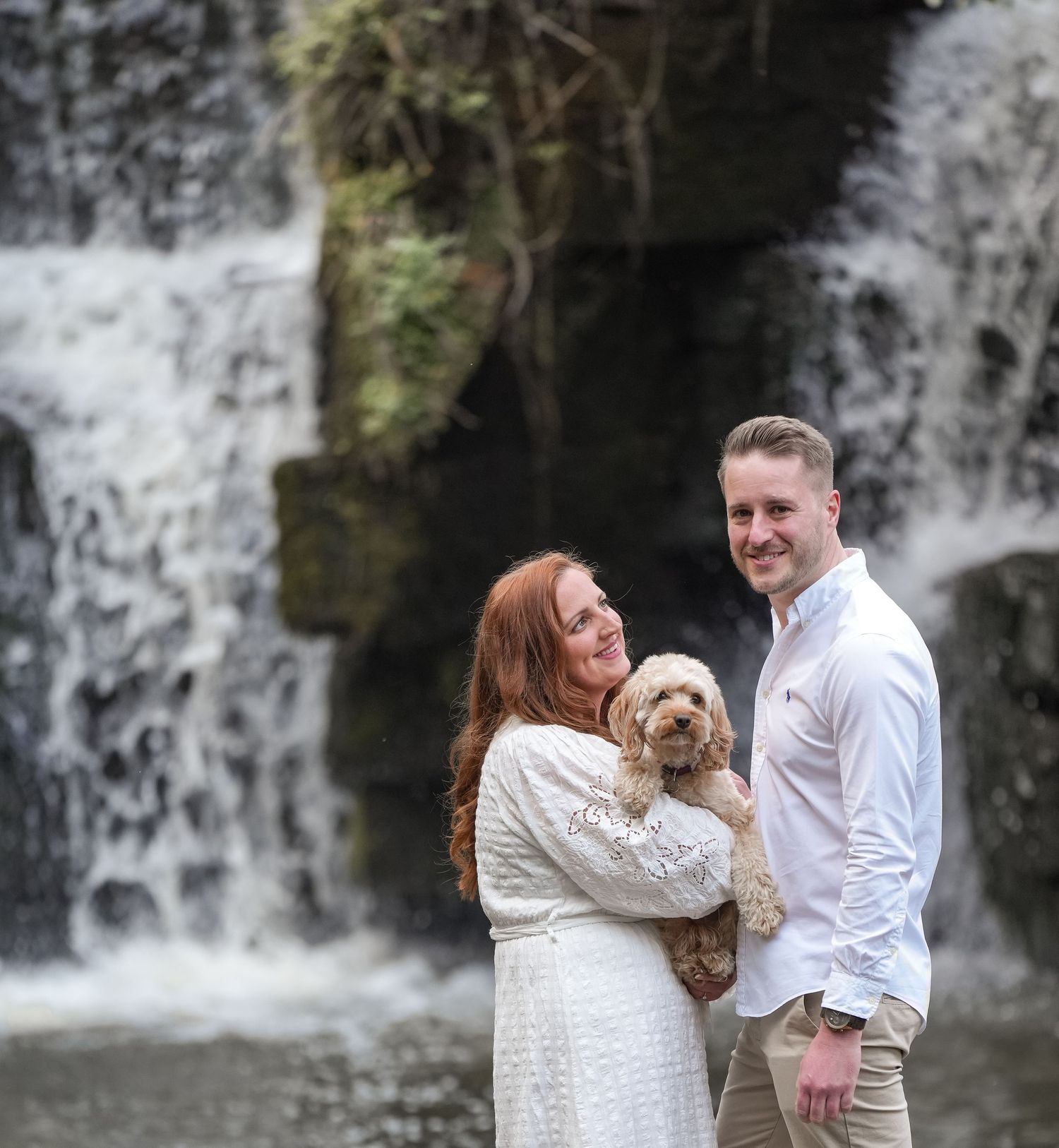 Couple poses with their dog near a scenic waterfall while dressed in white.