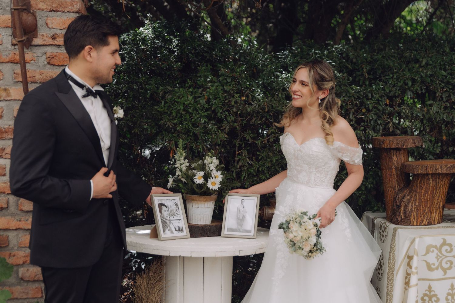 Bride and groom standing at decorated table with framed photos, exchanging smiles at outdoor wedding.