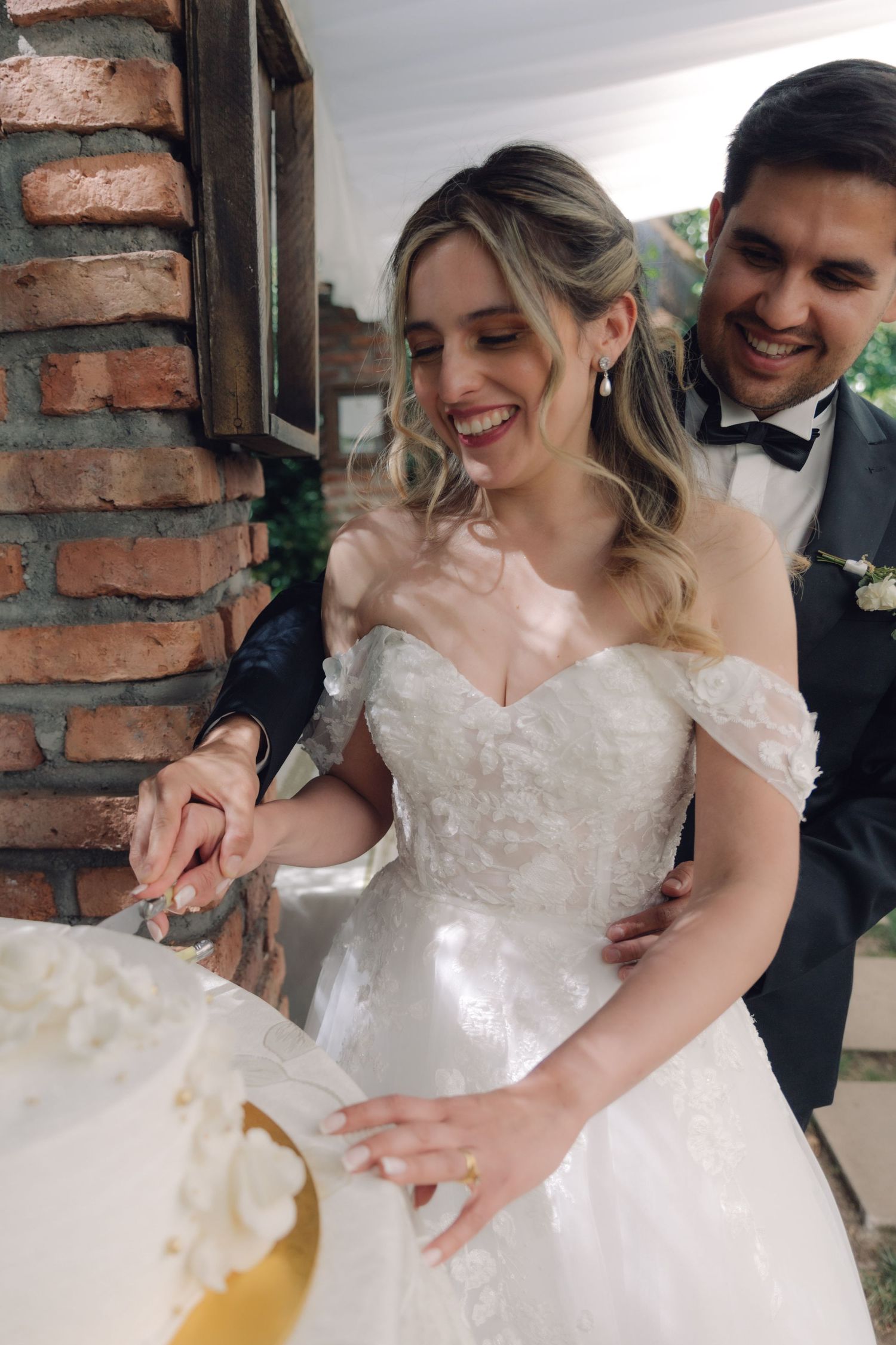Smiling bride and groom cutting white wedding cake together at outdoor brick-wall venue.