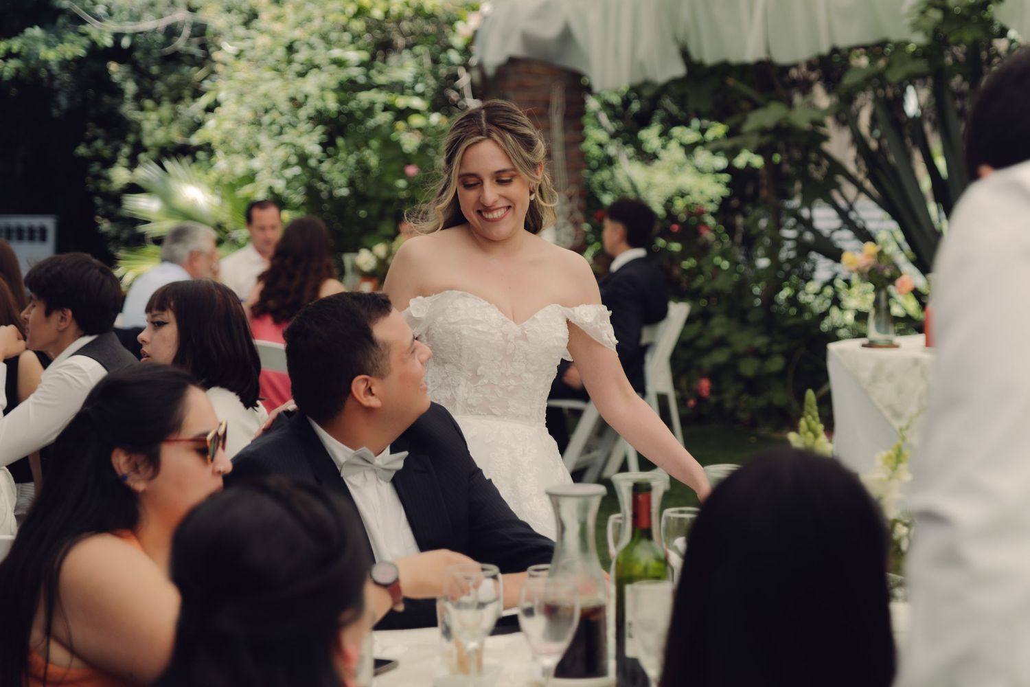 Radiant bride in strapless lace gown greeting seated wedding reception guests at outdoor venue.