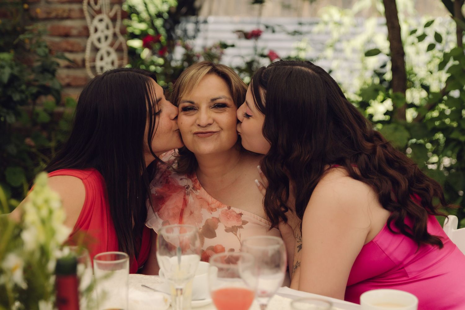 Two women kissing a smiling older woman on the cheeks at a festive wedding reception table.