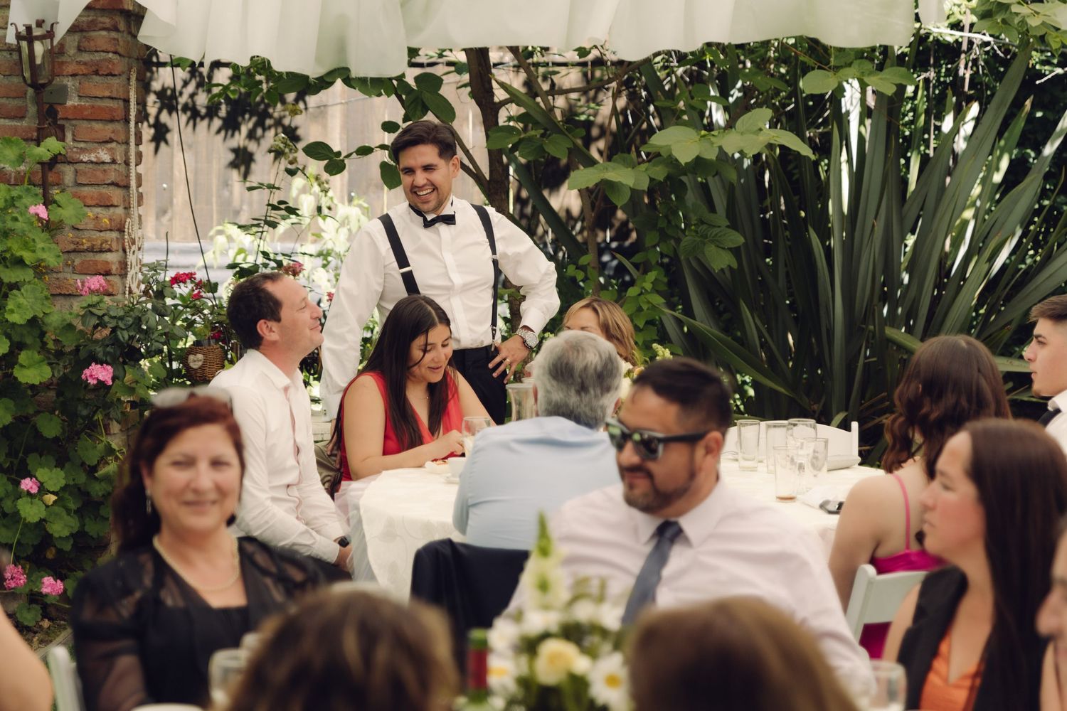 Groom in suspenders laughs with wedding guests at elegantly decorated outdoor garden reception.