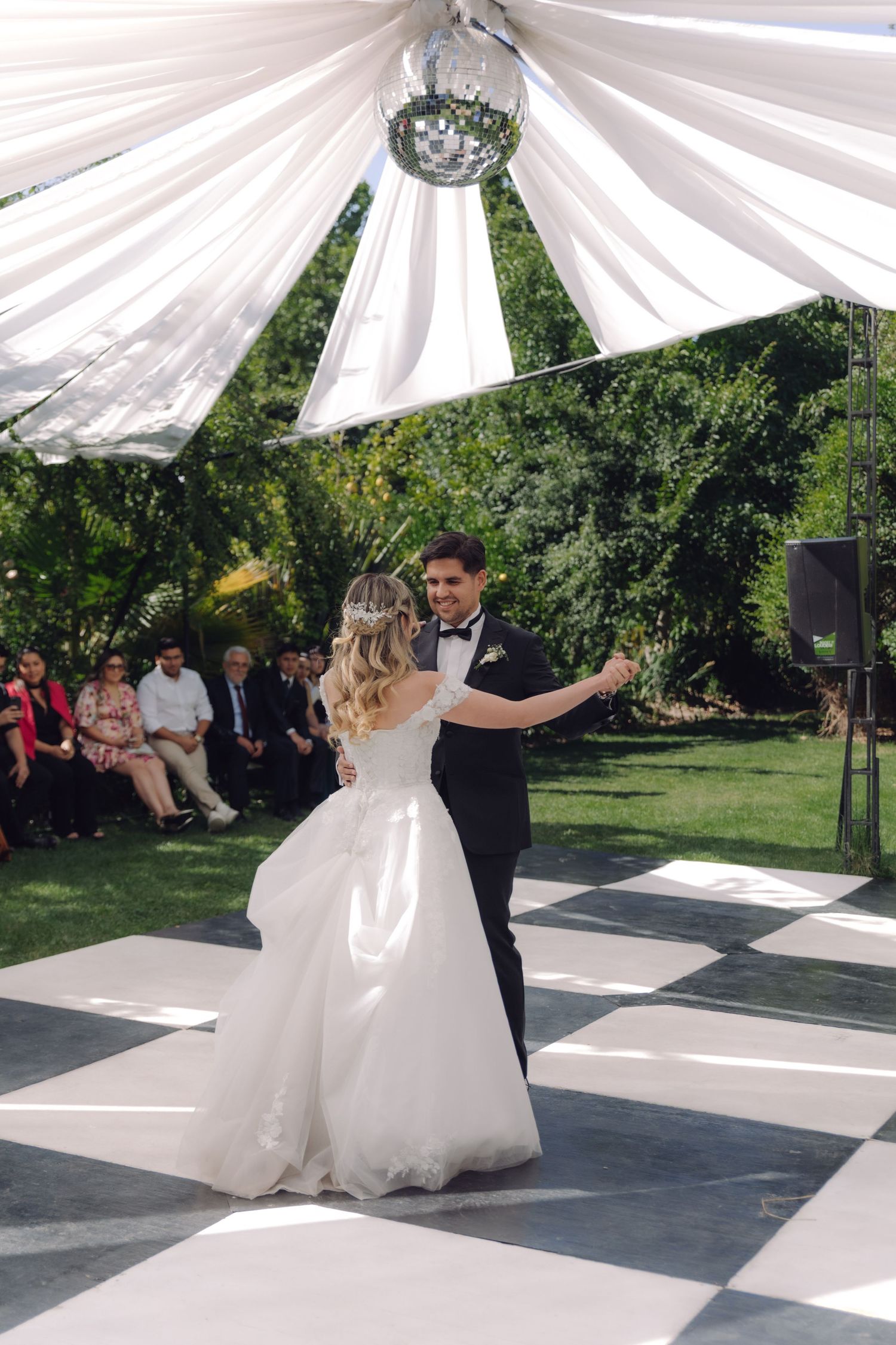 Bride and groom share their first dance on checkered floor under white draped canopy outdoors.