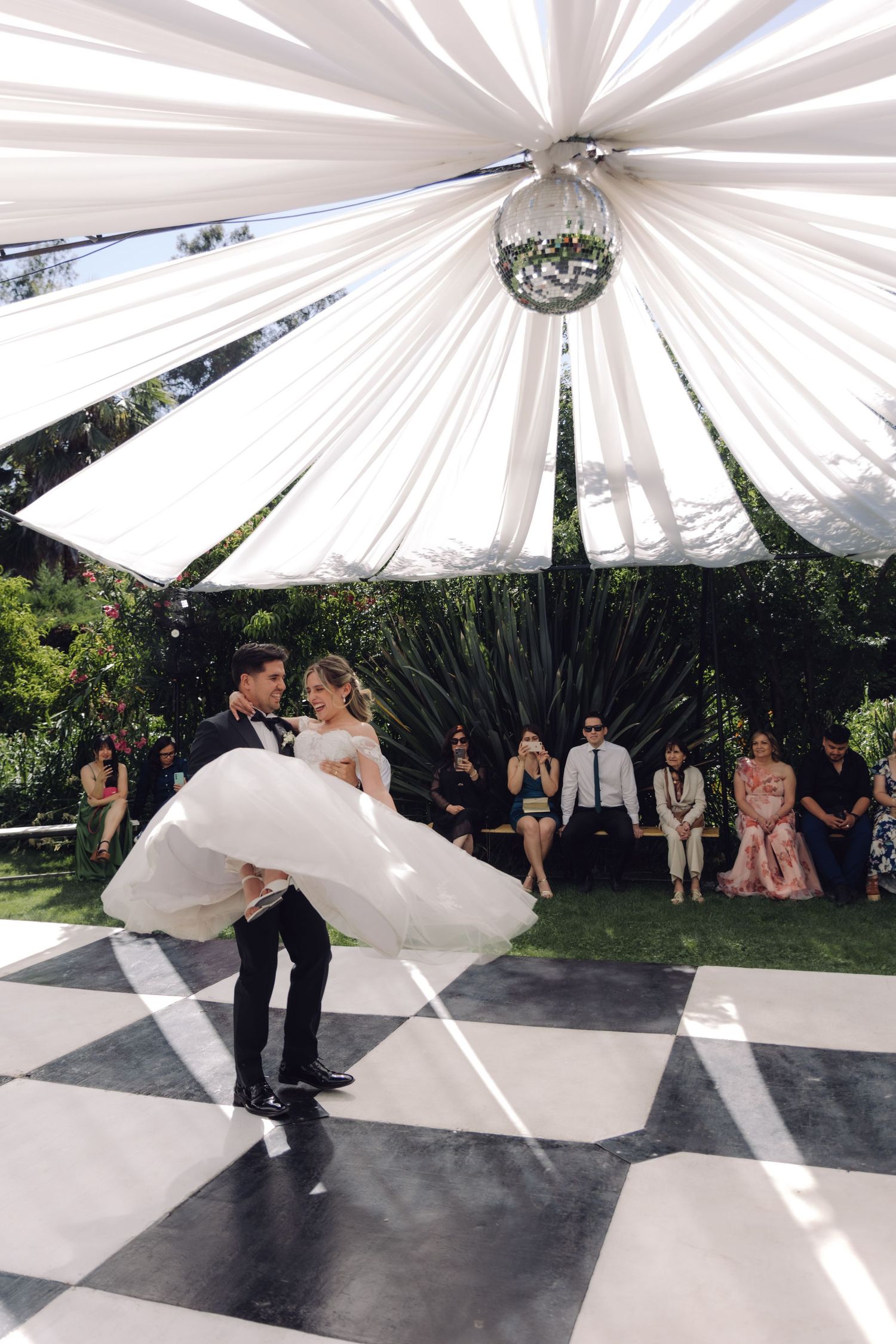 Groom twirls bride in flowing white gown on outdoor checkered dance floor under disco ball.