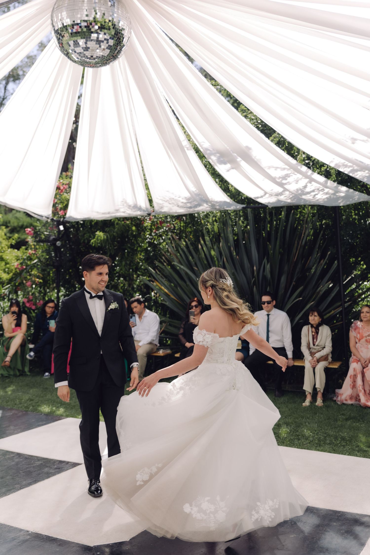 Bride in lace gown and groom in tuxedo dance elegantly on outdoor checkered wedding dance floor.