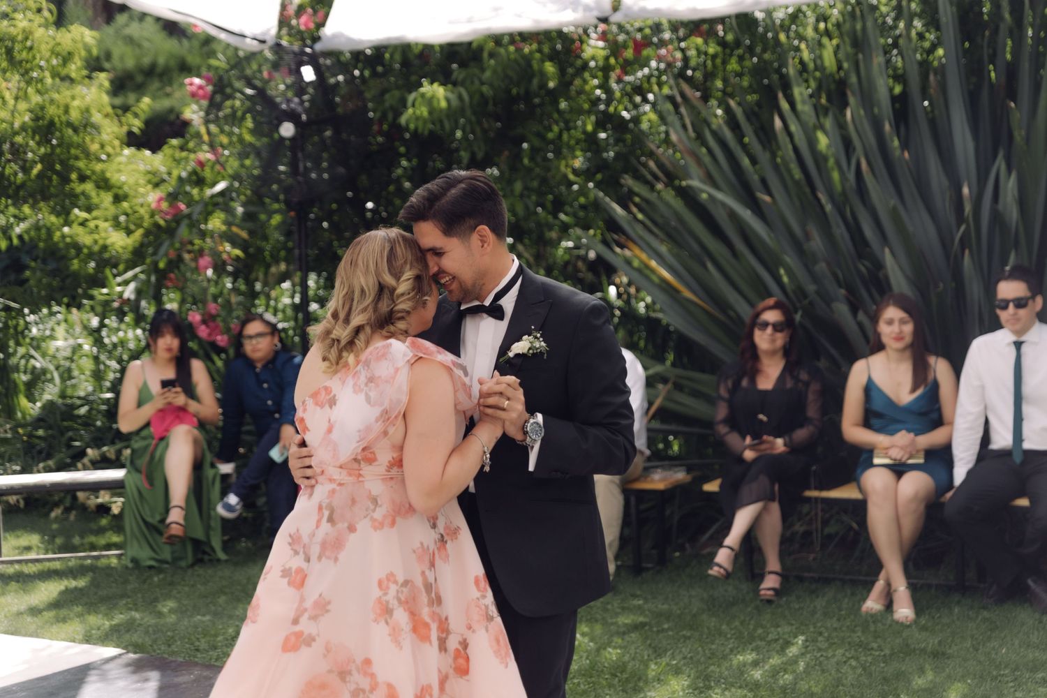 Couple shares a tender dance on outdoor dance floor as wedding guests watch in garden setting.