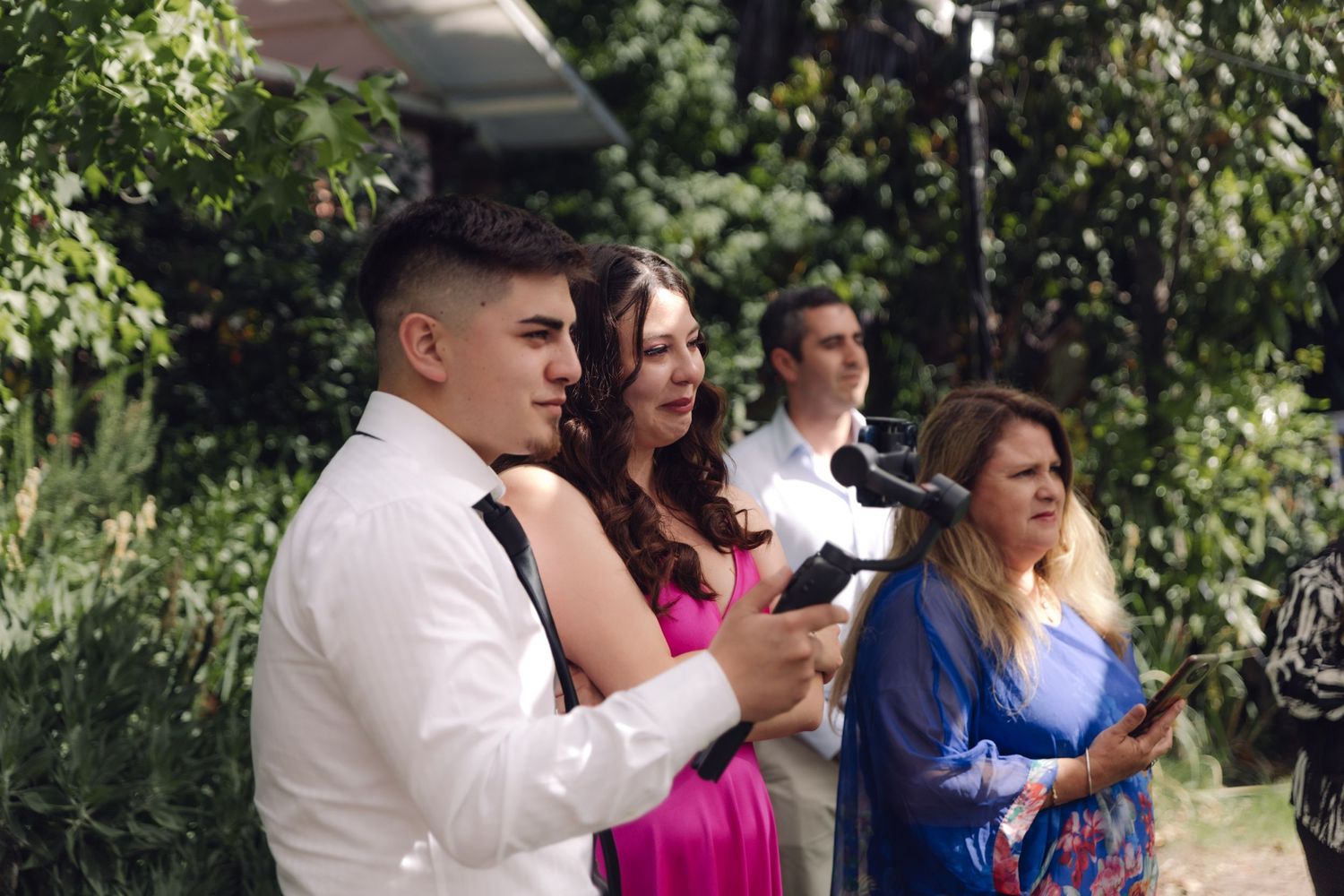 Young man and woman in formal attire speak at microphone during outdoor wedding celebration.