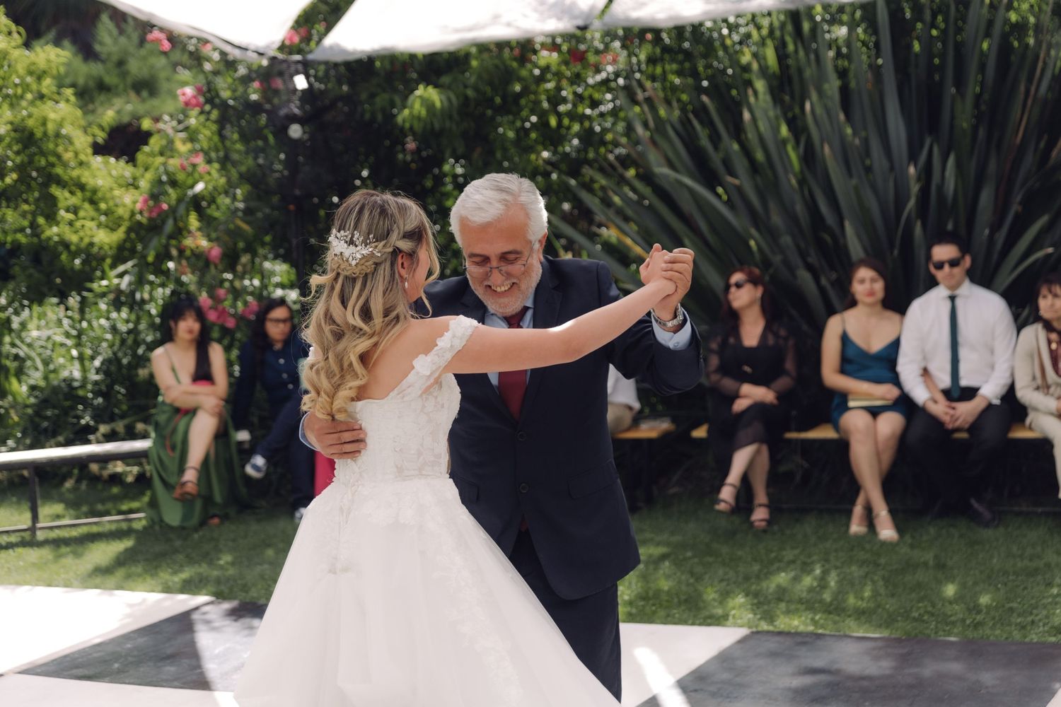 Bride in white gown dances with older gentleman in navy suit at outdoor garden wedding.
