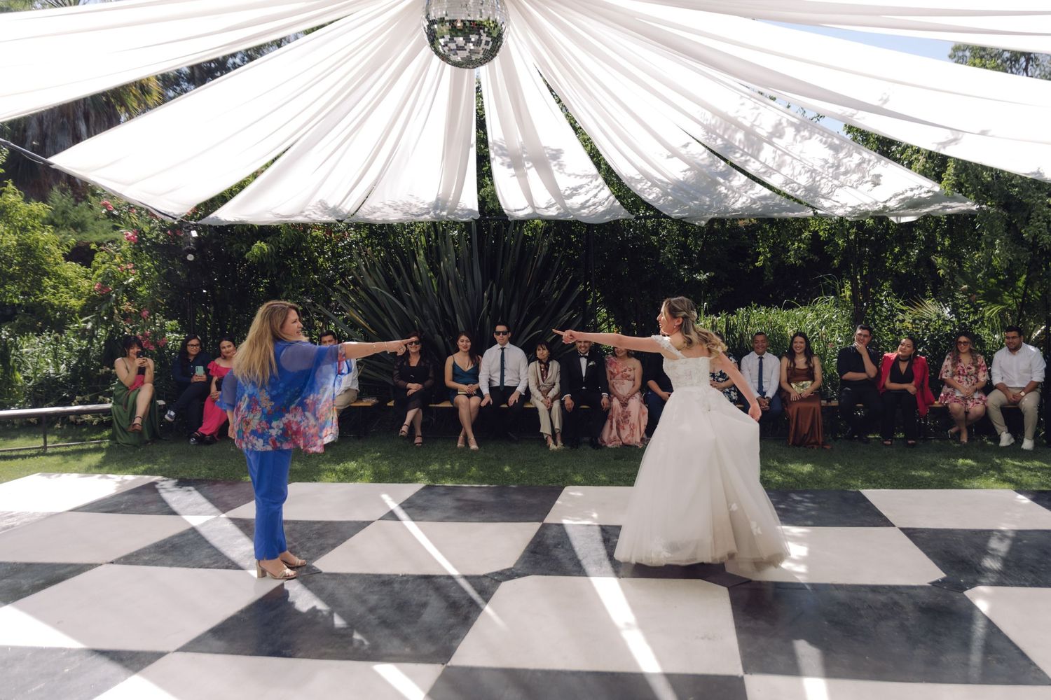 Bride and guest dancing together on checkered dance floor at outdoor wedding reception under white draped tent.