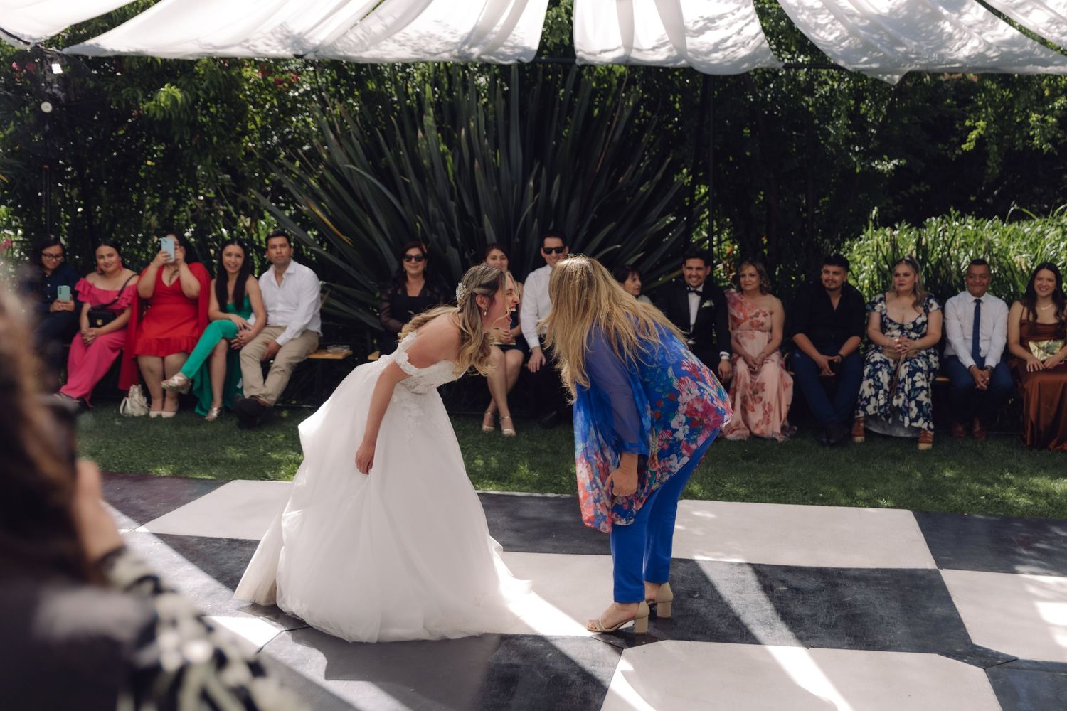Bride in white gown bowing with guest on checkered dance floor at outdoor wedding reception.