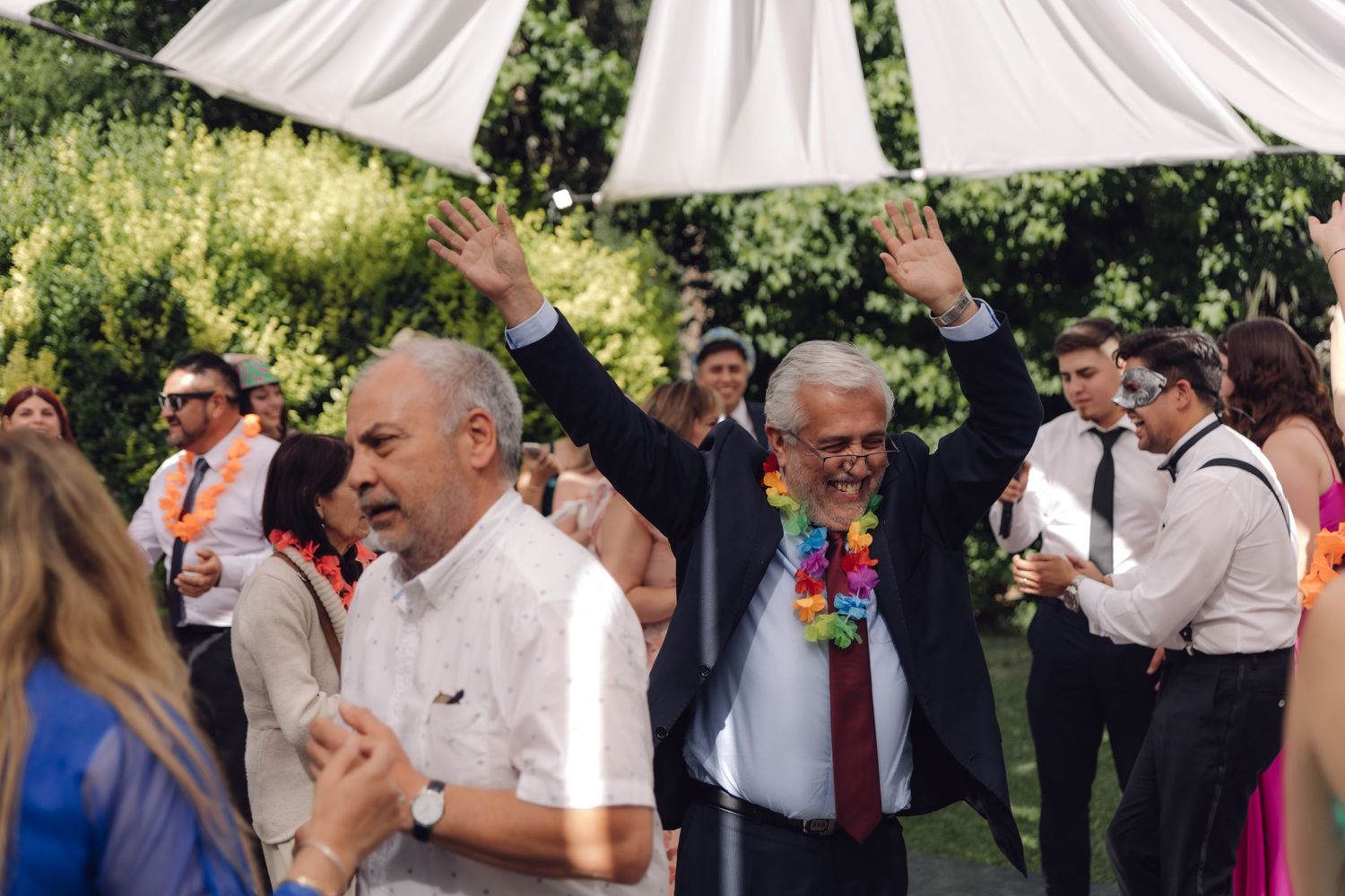 Two men celebrating at outdoor wedding reception, one wearing colorful Hawaiian lei with hands raised.