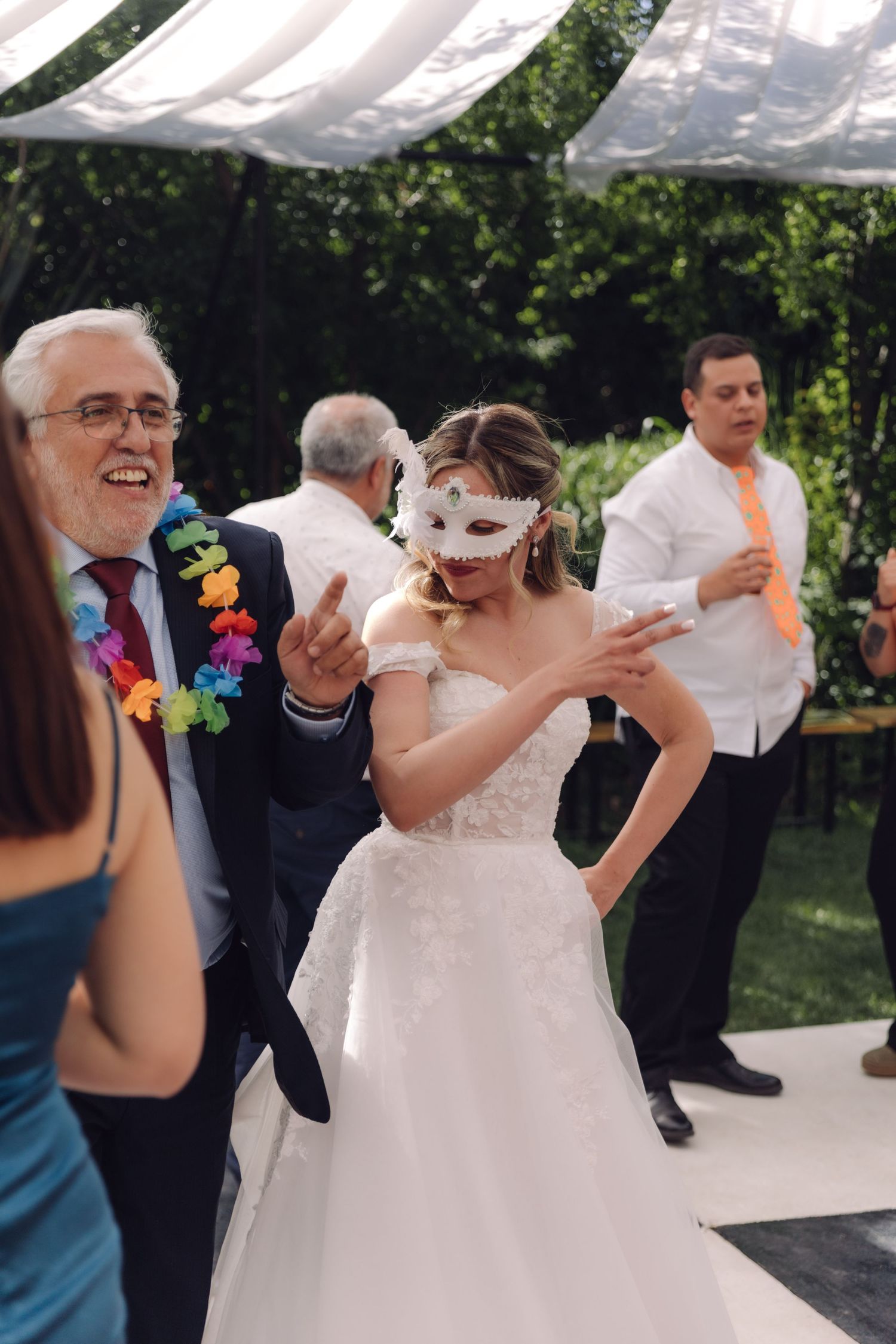 Bride wearing white masquerade mask dancing with older man in suit at outdoor wedding reception.