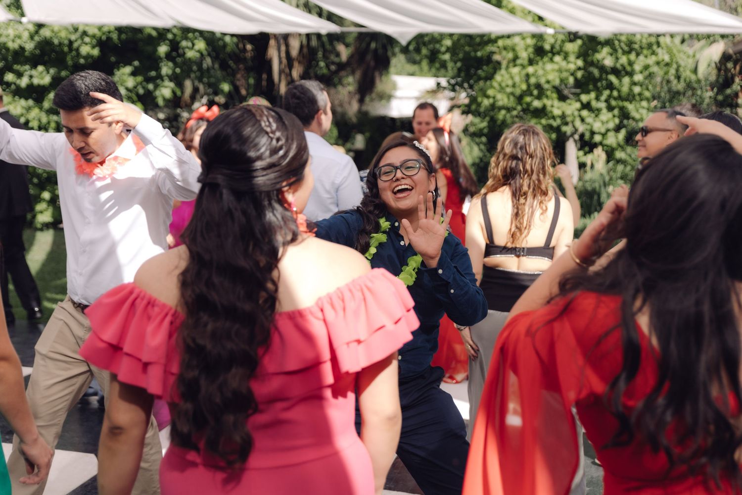 Guests dancing joyfully at outdoor wedding reception on sunny day under white tent canopy.