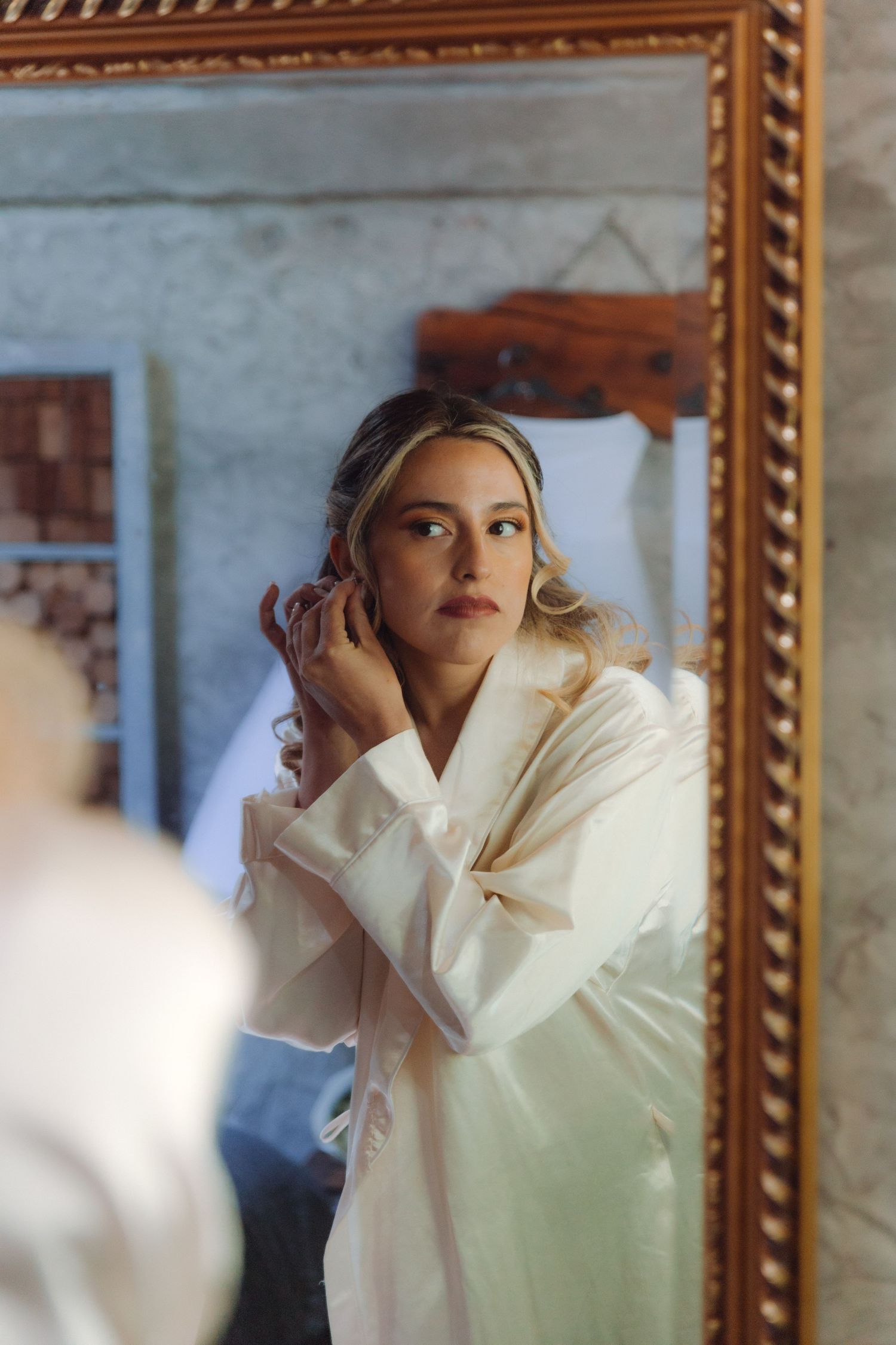 Bride in white robe adjusting earrings while looking into an ornate gold-framed mirror.