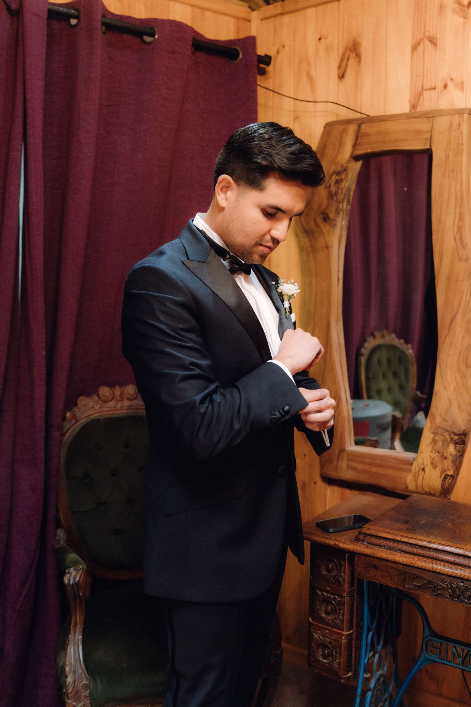 Groom in black tuxedo adjusting boutonniere while standing near a wooden mirror indoors.