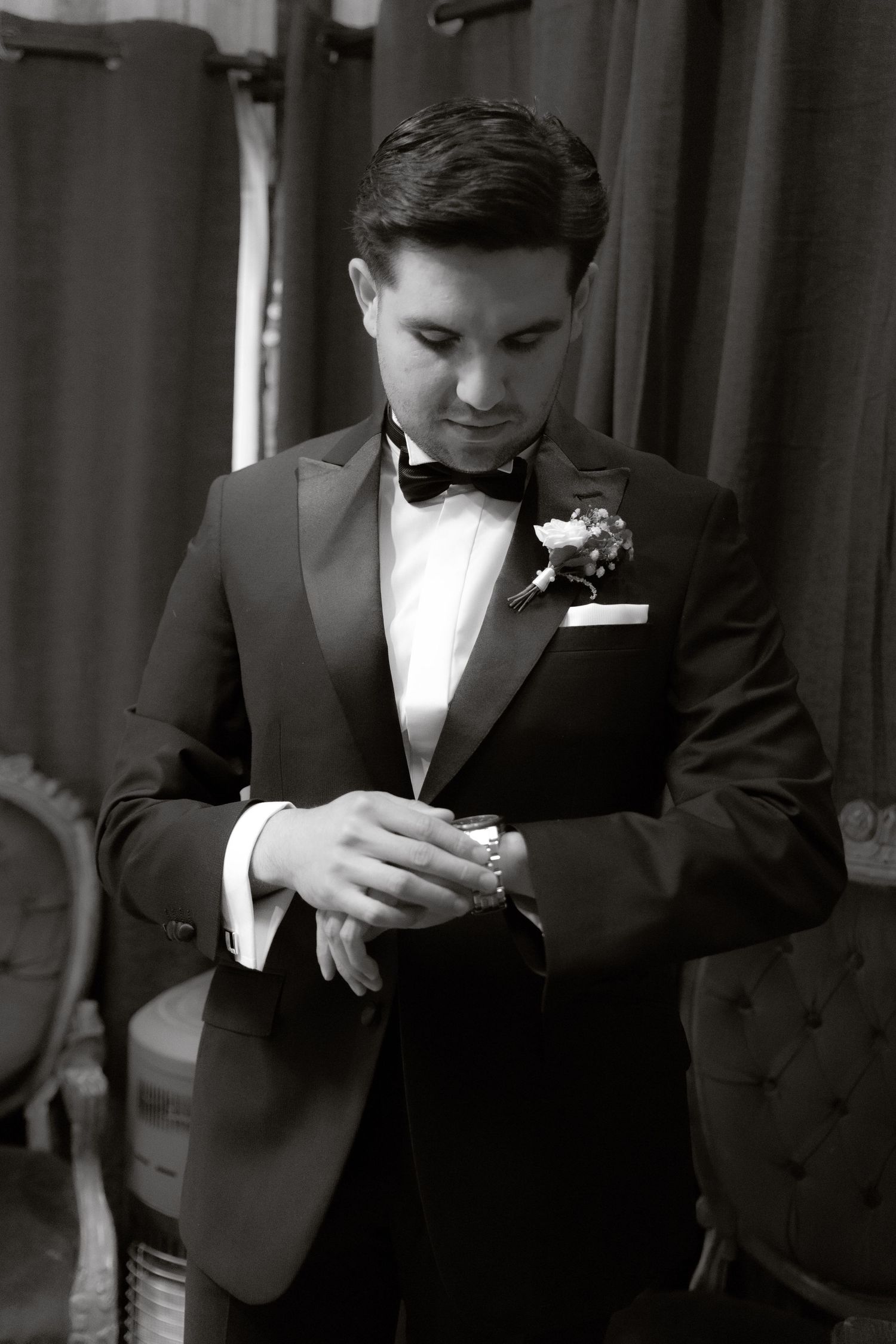 Black and white portrait of a groom in a tuxedo with boutonniere adjusting his cufflinks.