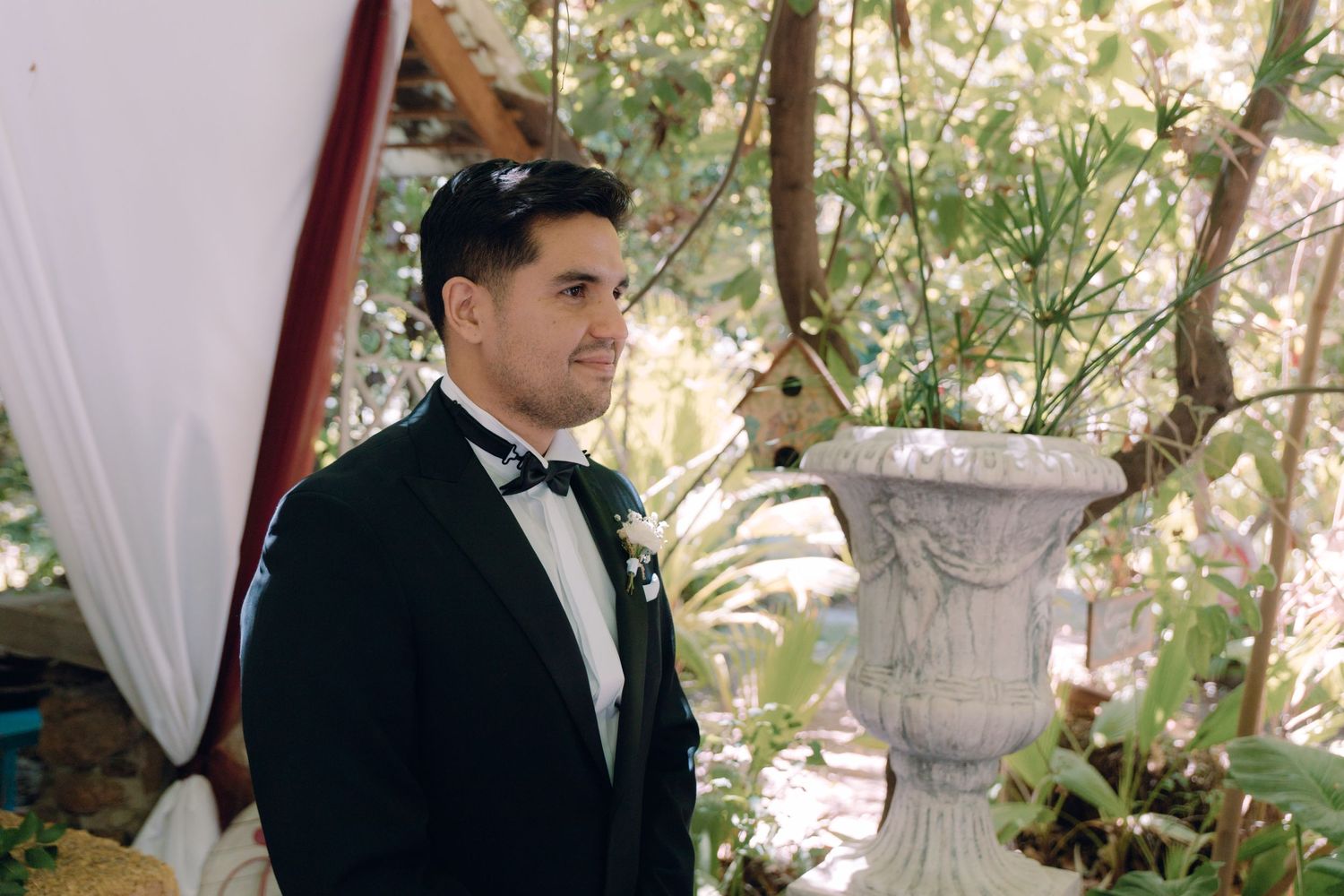 Groom in black tuxedo with bow tie stands at outdoor wedding altar surrounded by lush greenery.