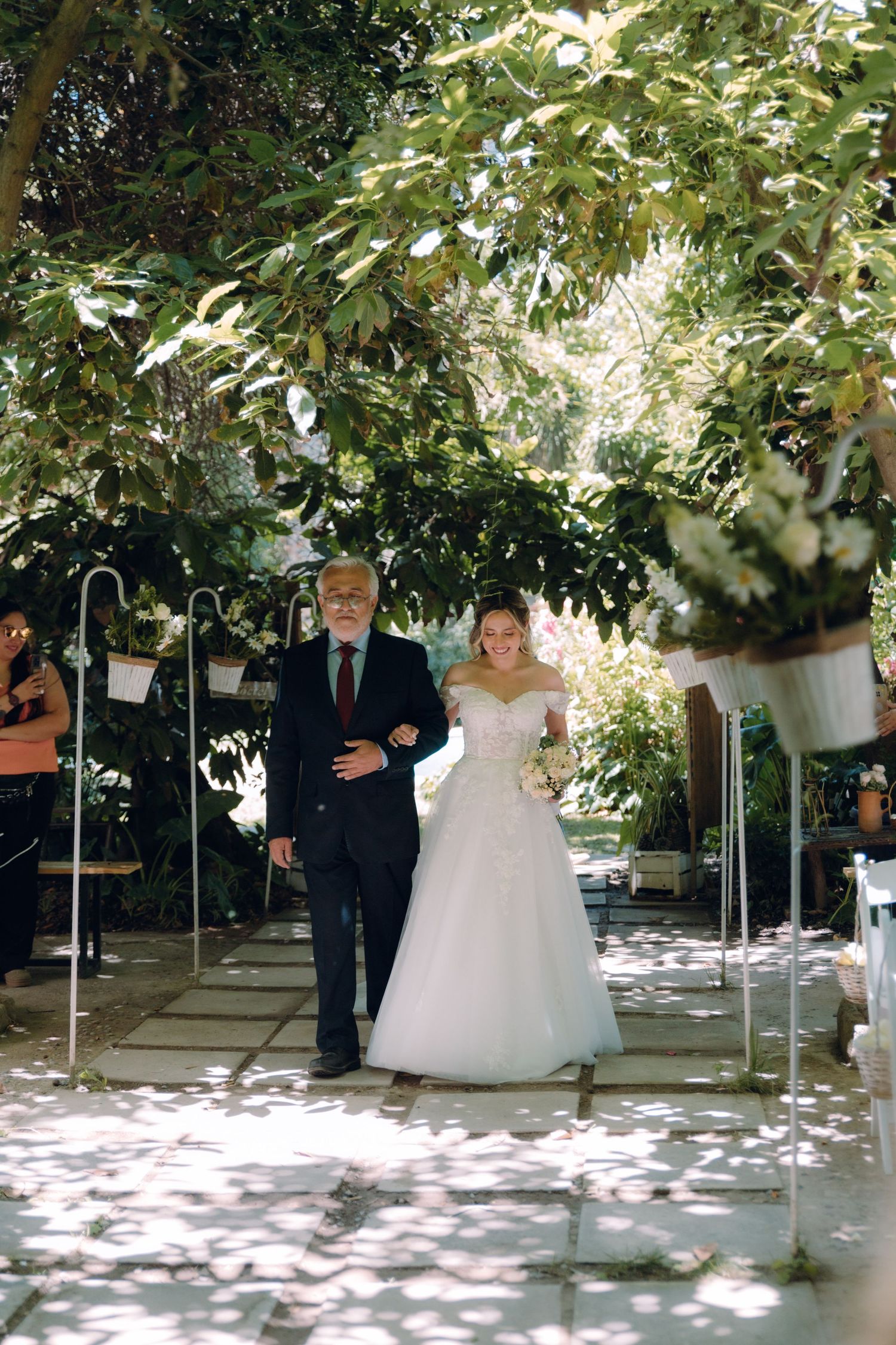 Bride in white off-shoulder gown walks down aisle with older gentleman escort at outdoor garden wedding.