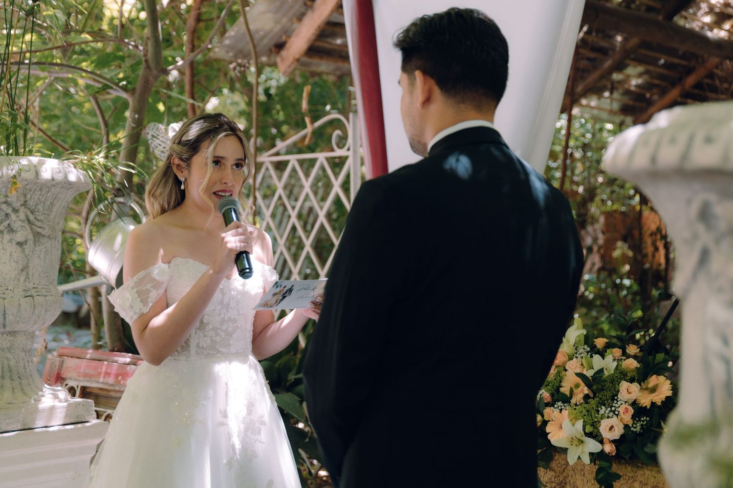 Bride in white dress reads wedding vows into microphone at romantic outdoor garden ceremony.