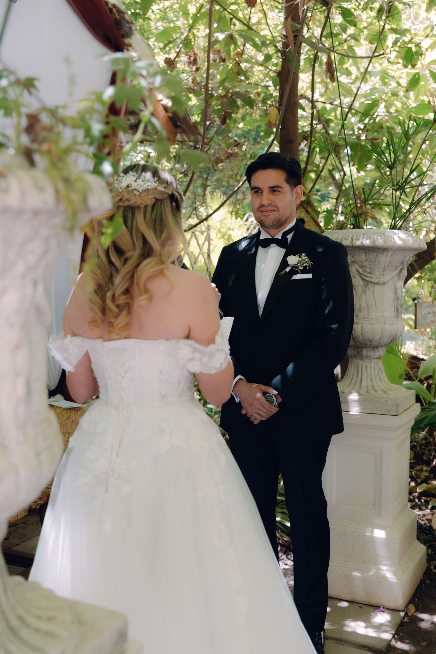 Groom in black tuxedo gazes at bride in white lace gown during outdoor garden wedding ceremony.