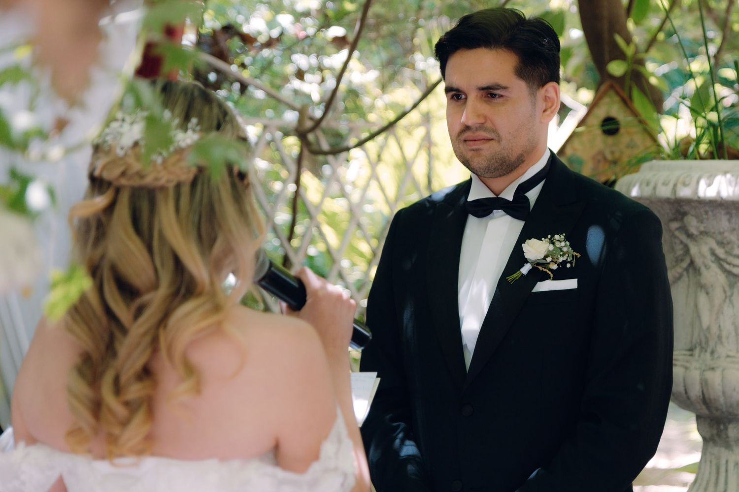 Groom in black tuxedo listens attentively as bride recites wedding vows at outdoor ceremony.