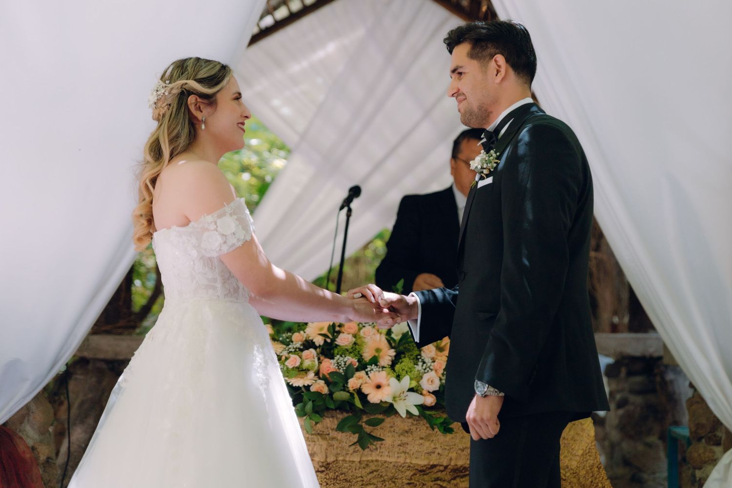 Bride and groom exchange rings at outdoor wedding altar decorated with pink and white flowers.