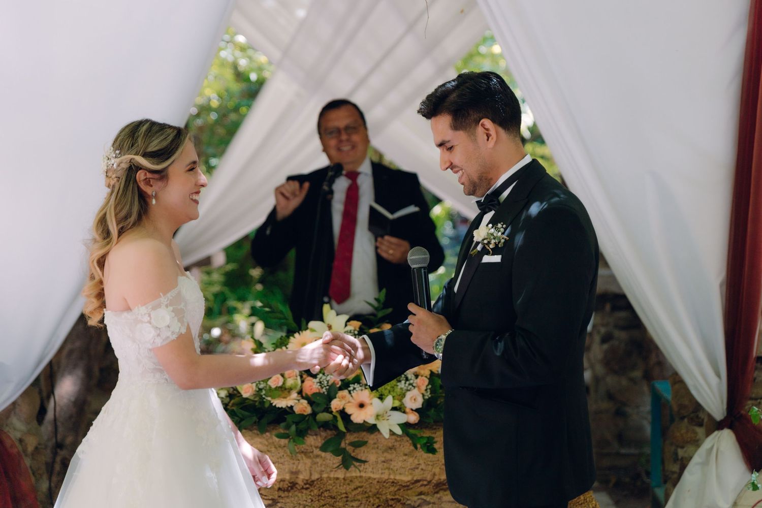 Smiling bride and groom exchange vows with officiant at floral-decorated outdoor wedding ceremony.