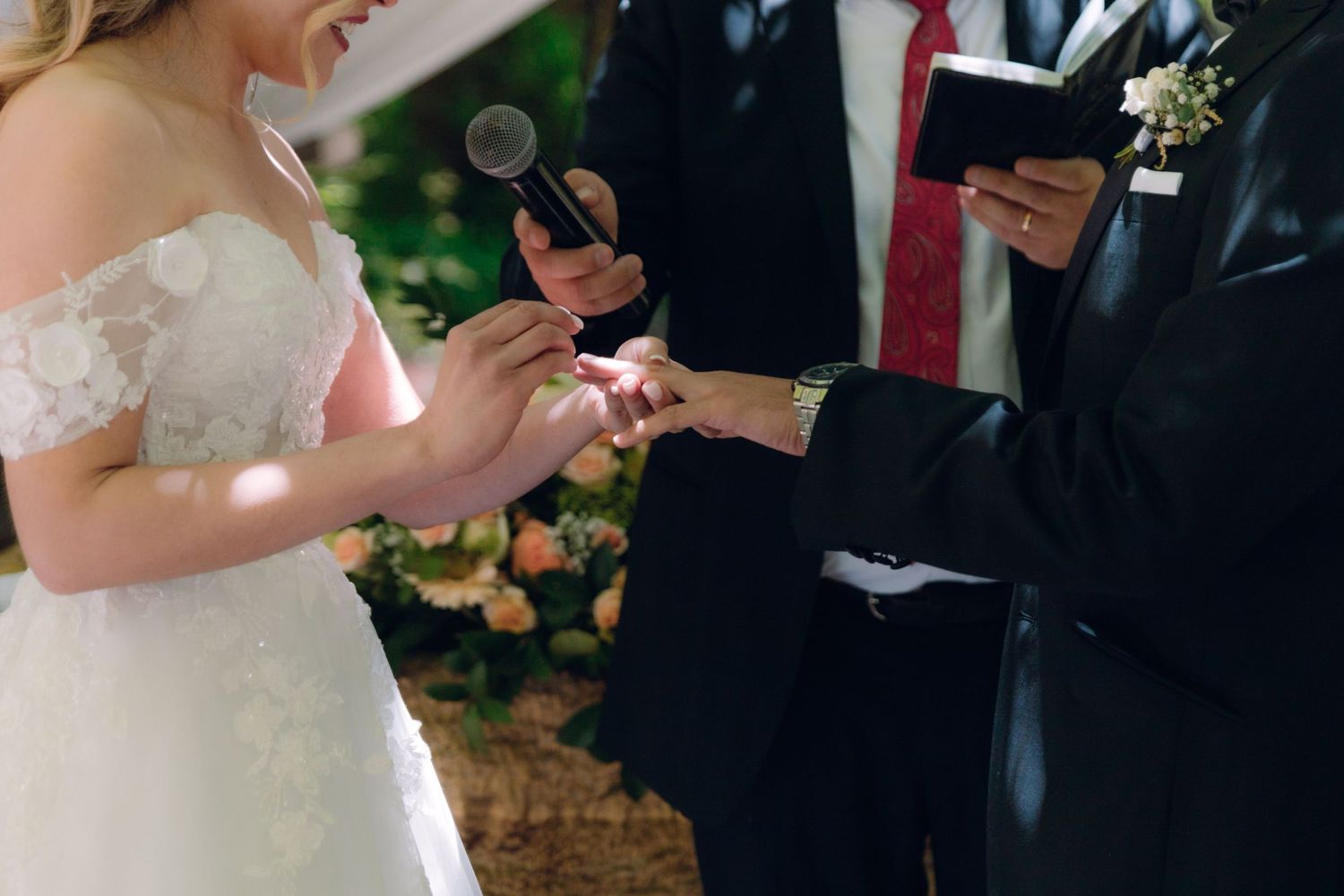 Close-up of bride placing wedding ring on grooms finger during outdoor ceremony with floral backdrop.