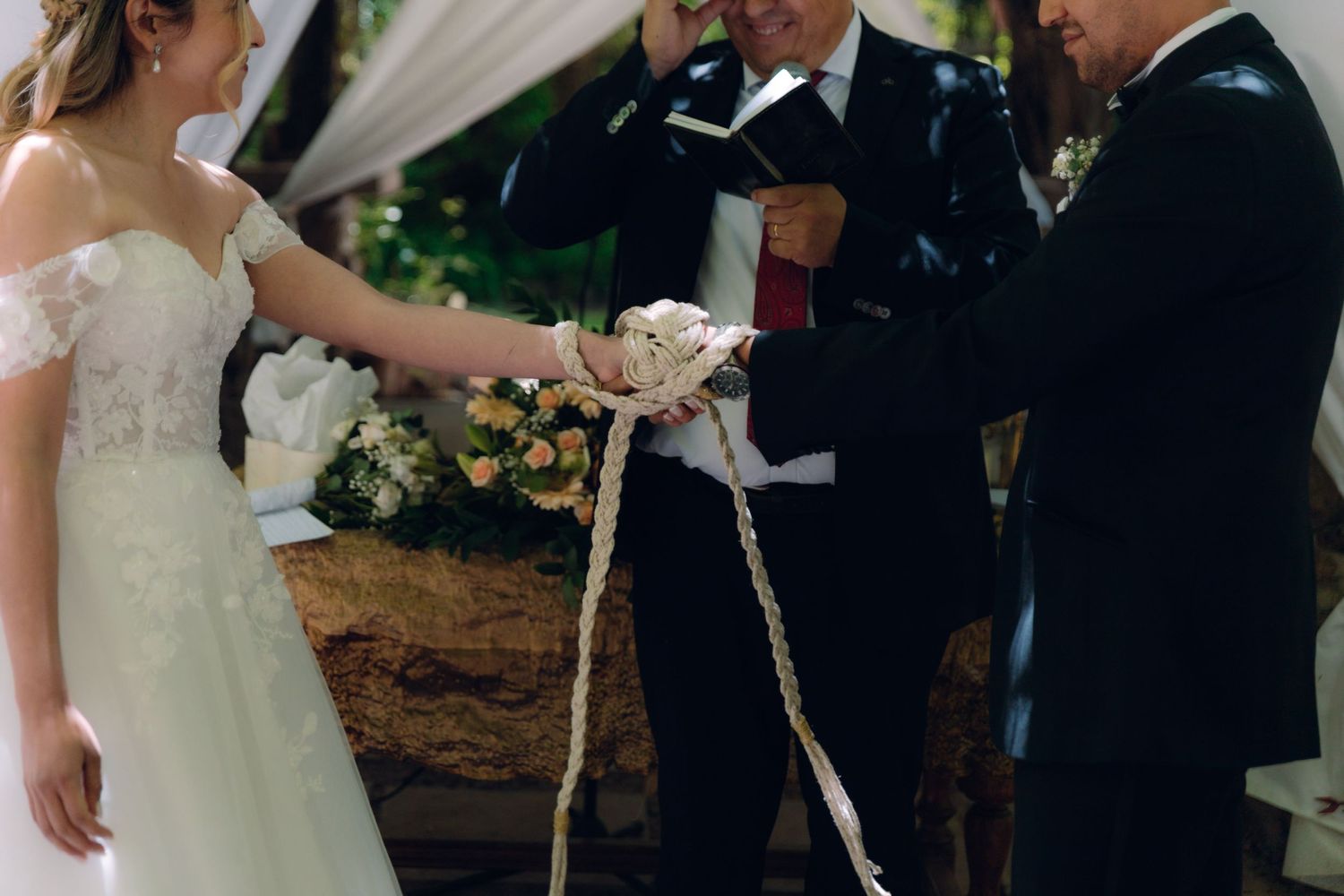 Bride and groom participate in rope unity ceremony with officiant at outdoor garden wedding.