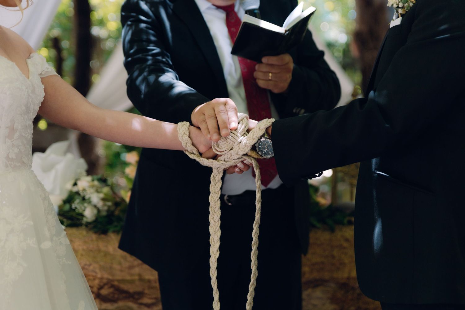Officiant performing handfasting ceremony tying rope around bride and groom hands at outdoor wedding.
