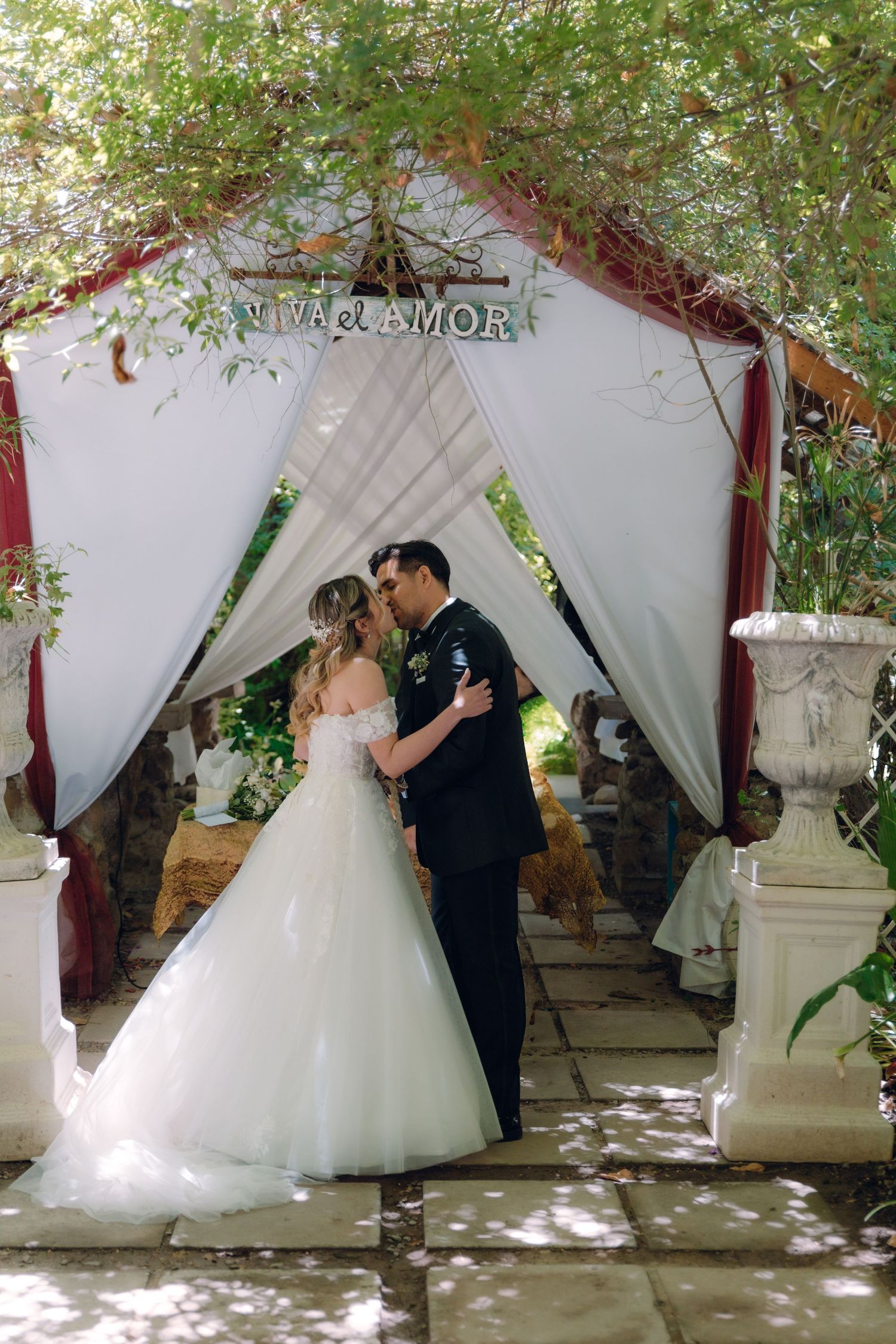 Bride and groom share first kiss under floral arch with Viva el Amor sign at outdoor wedding.