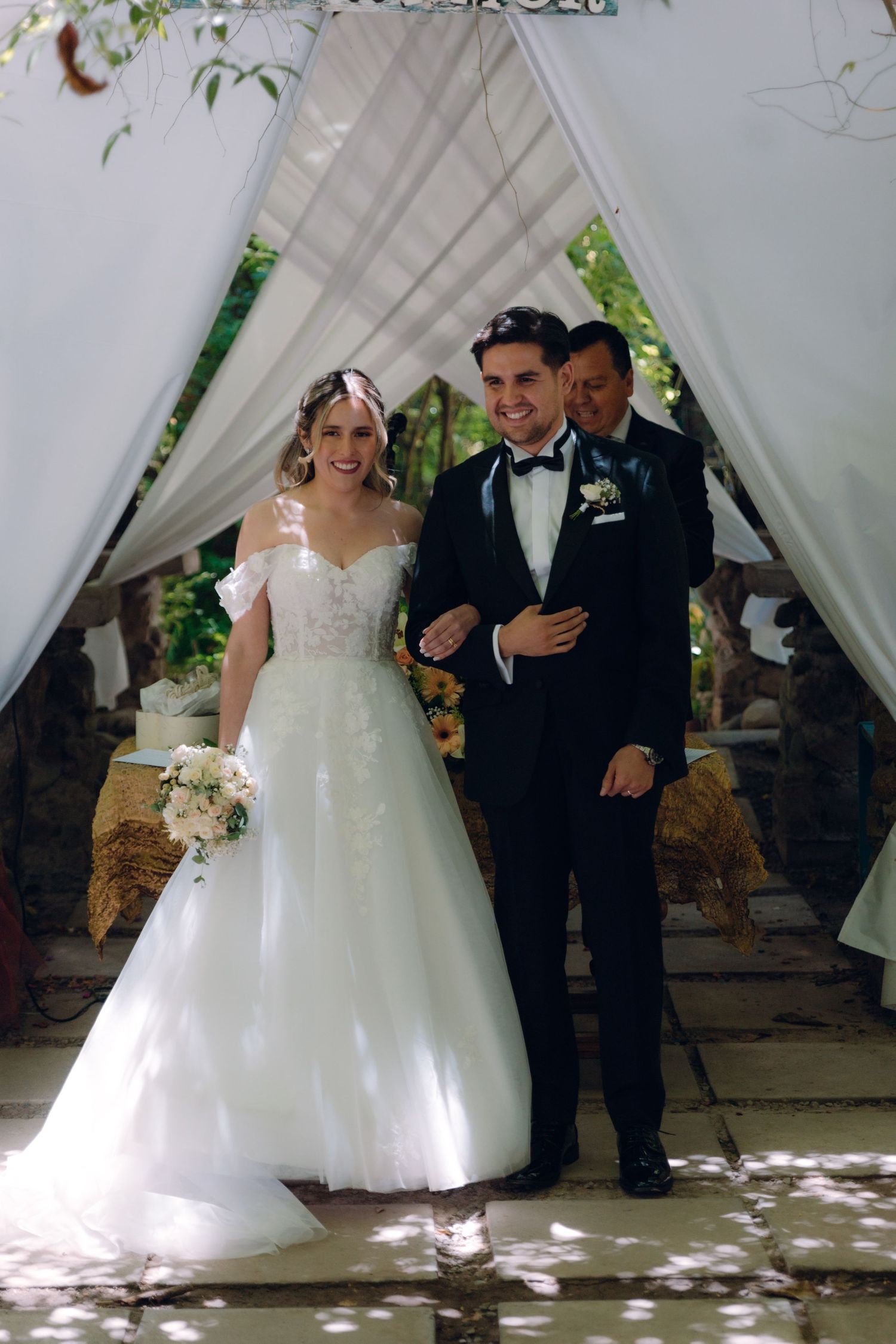 Smiling bride in lace off-shoulder gown walks with groom in tuxedo under wedding ceremony arch.