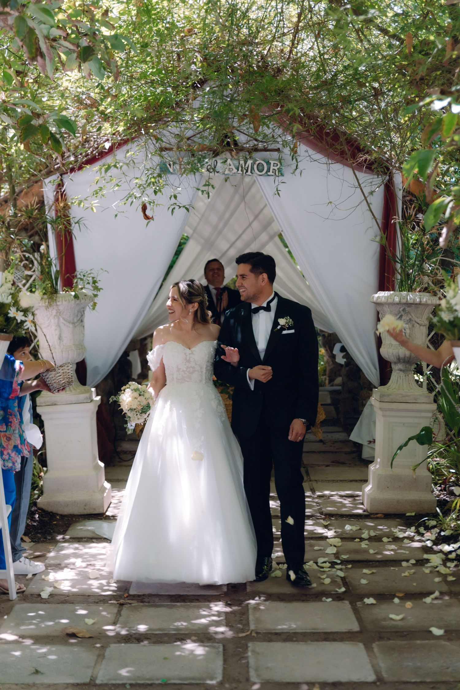 Newlyweds walk down aisle under decorated arch with Viva el Amor sign at outdoor garden wedding.