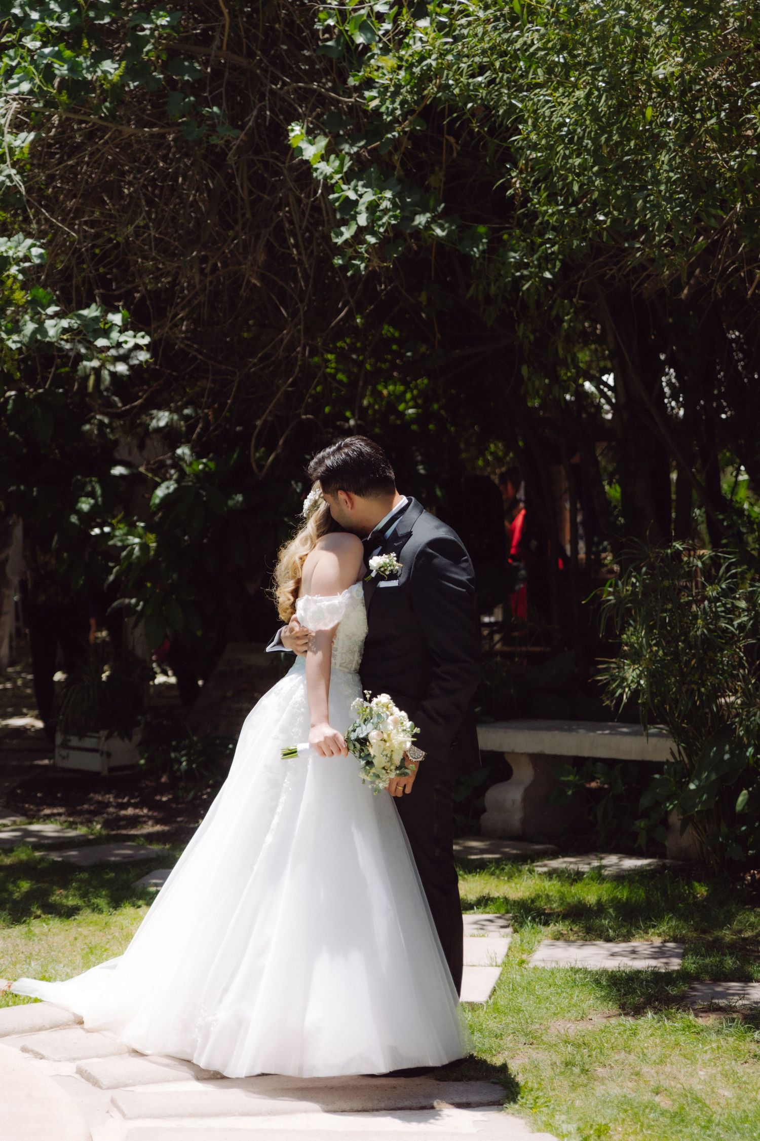 Bride and groom share romantic kiss in sunlit garden surrounded by lush green trees.