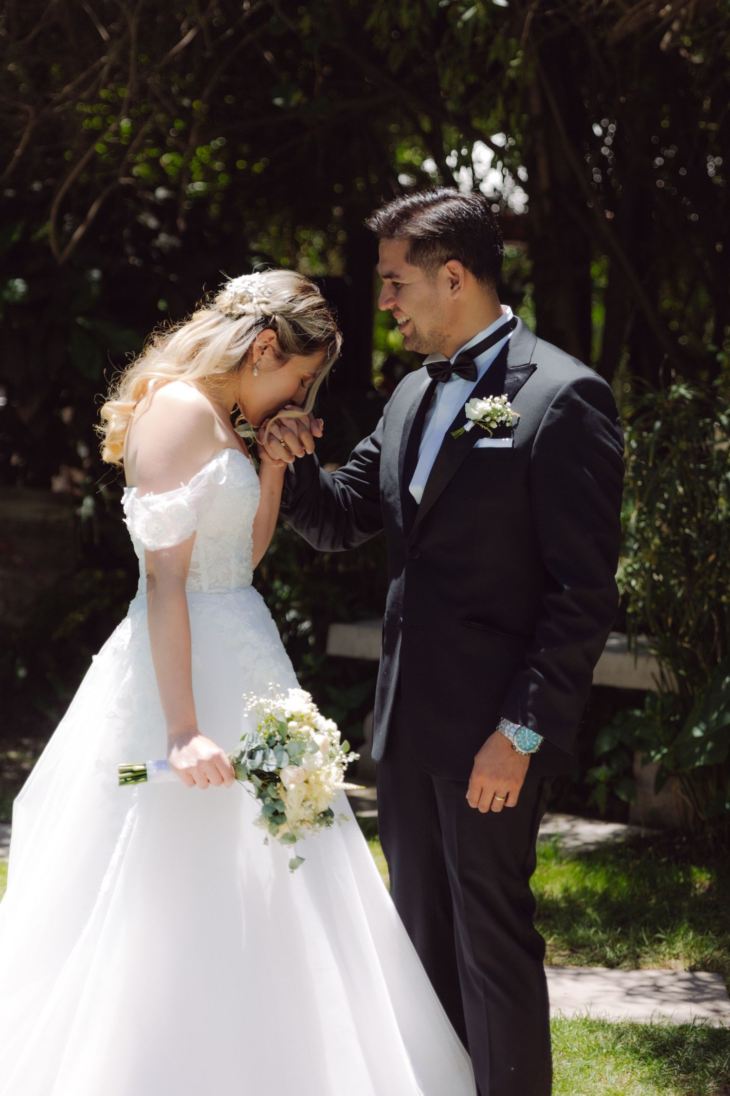 Groom in tuxedo kisses brides hand as she laughs holding white bouquet in garden setting.