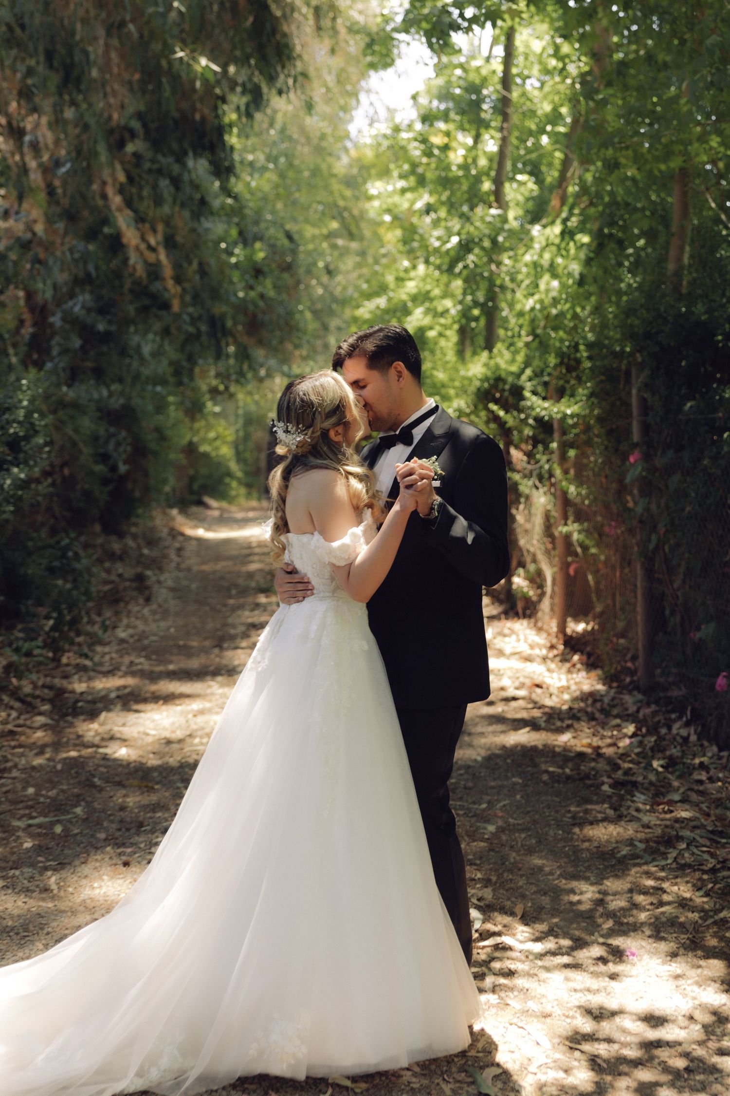 Bride and groom embrace tenderly on tree-lined garden path in dappled sunlight on wedding day.