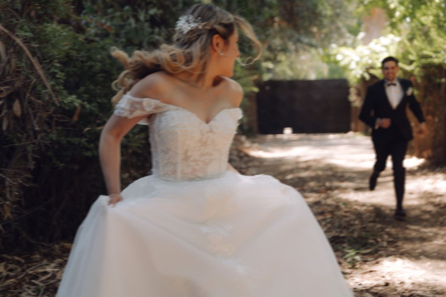 Bride in lace off-shoulder ball gown walks through garden path with groom approaching behind.
