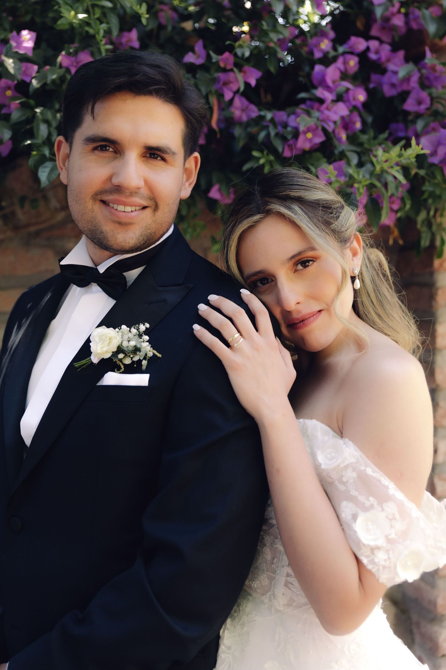 Bride and groom pose together smiling in front of purple flowering wall on wedding day.