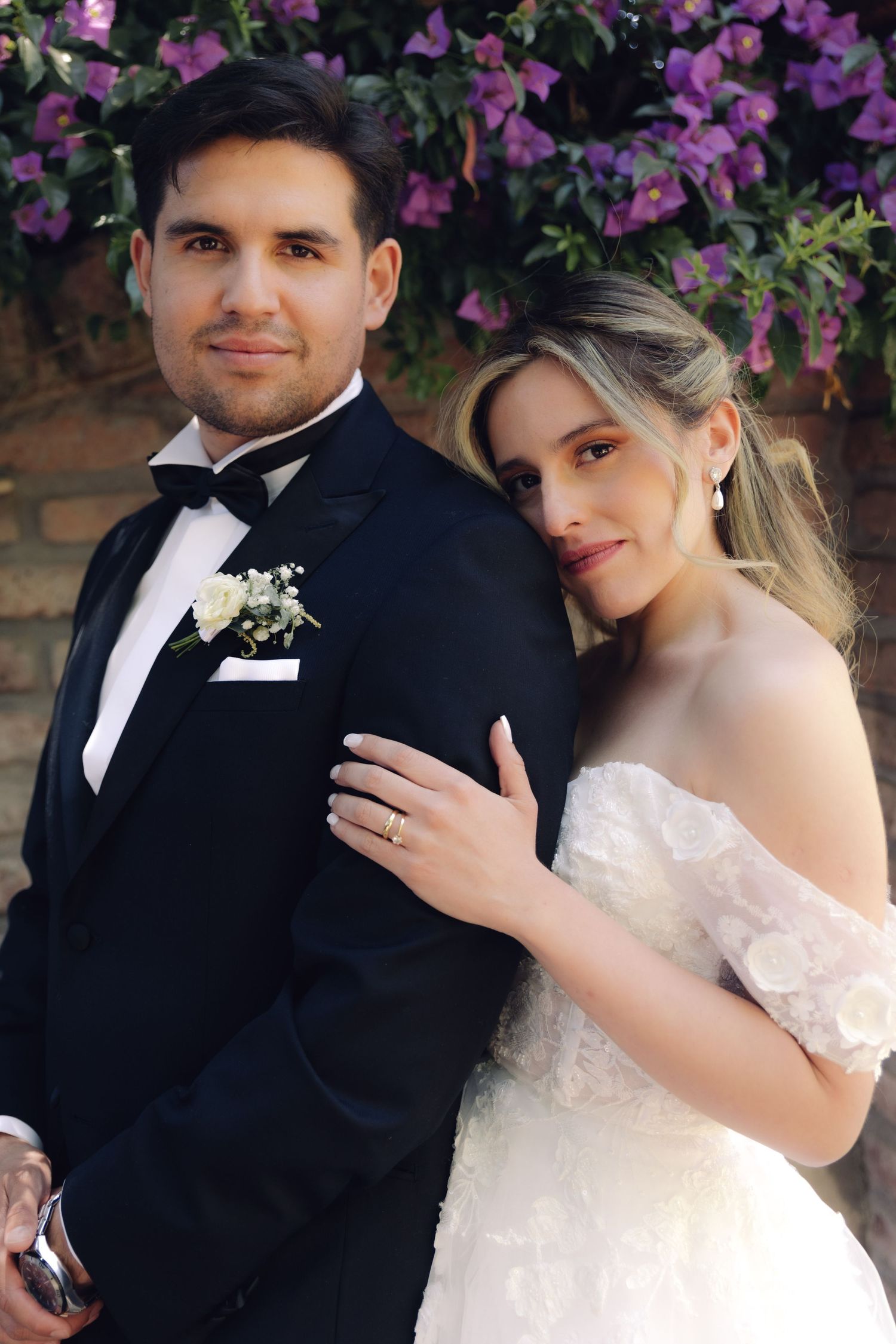 Bride and groom posing together in front of purple bougainvillea, wearing wedding attire with boutonniere.