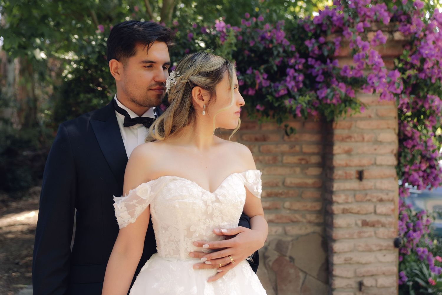 Newlyweds embracing outdoors near purple flowering arch, bride in off-shoulder lace gown and groom in tuxedo.