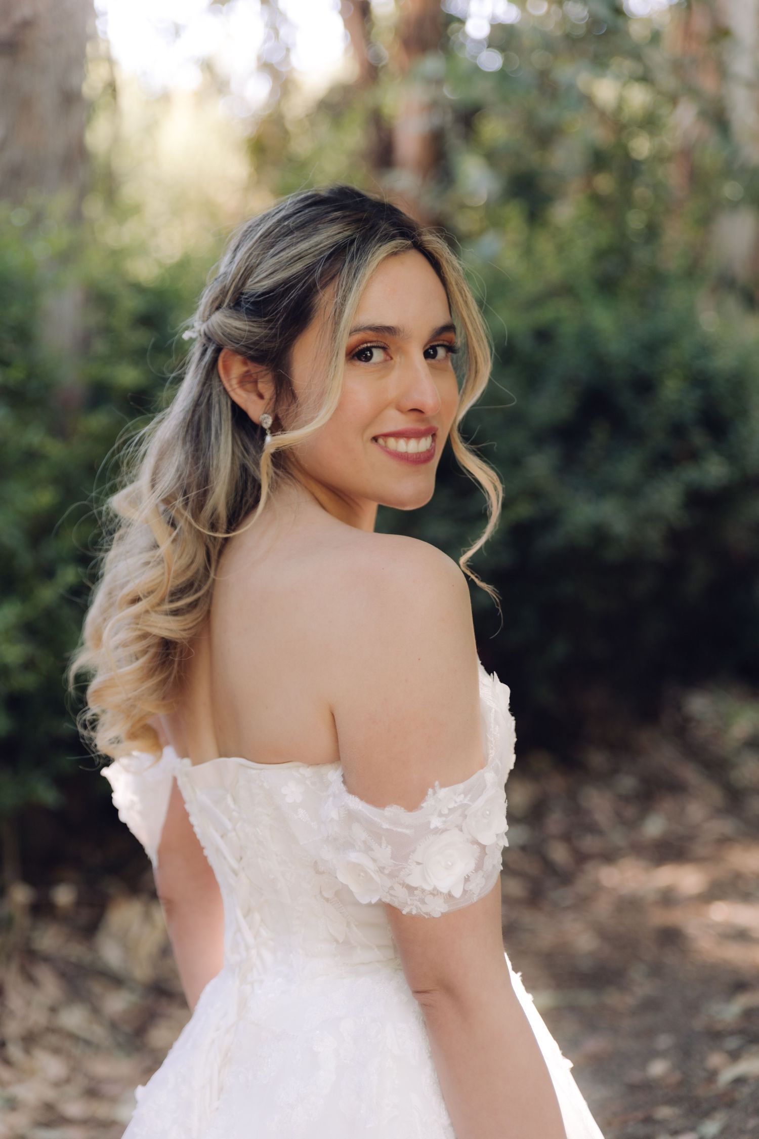 Smiling bride in off-shoulder lace wedding dress posing outdoors with soft natural lighting.