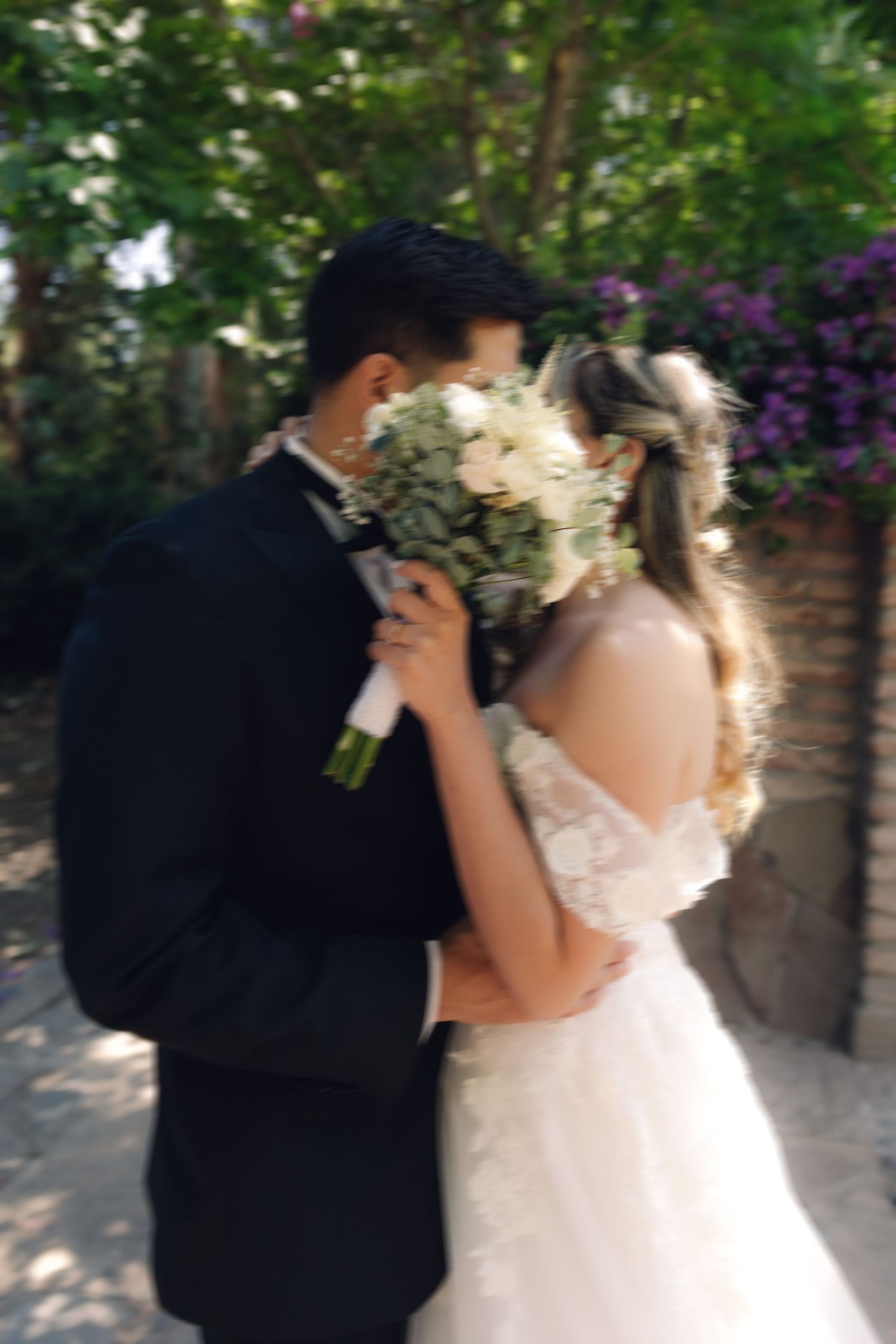 Bride and groom sharing a kiss hidden behind a white floral bouquet in a garden setting.