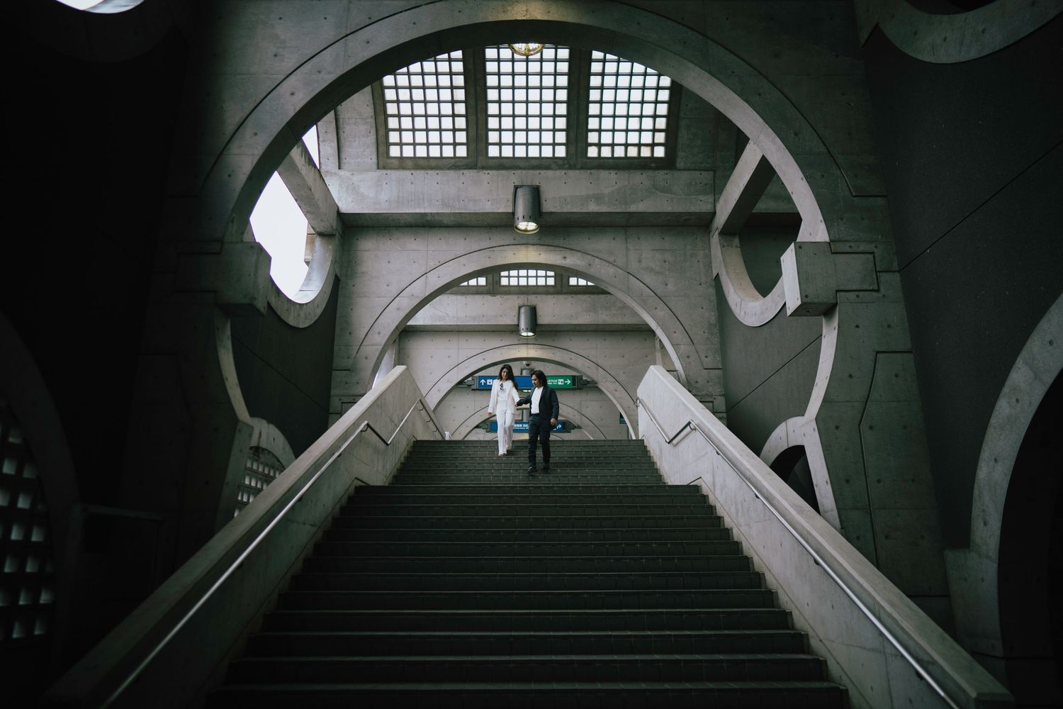 Couple walking up grand arched staircase inside a dramatic architectural building with high vaulted ceilings.