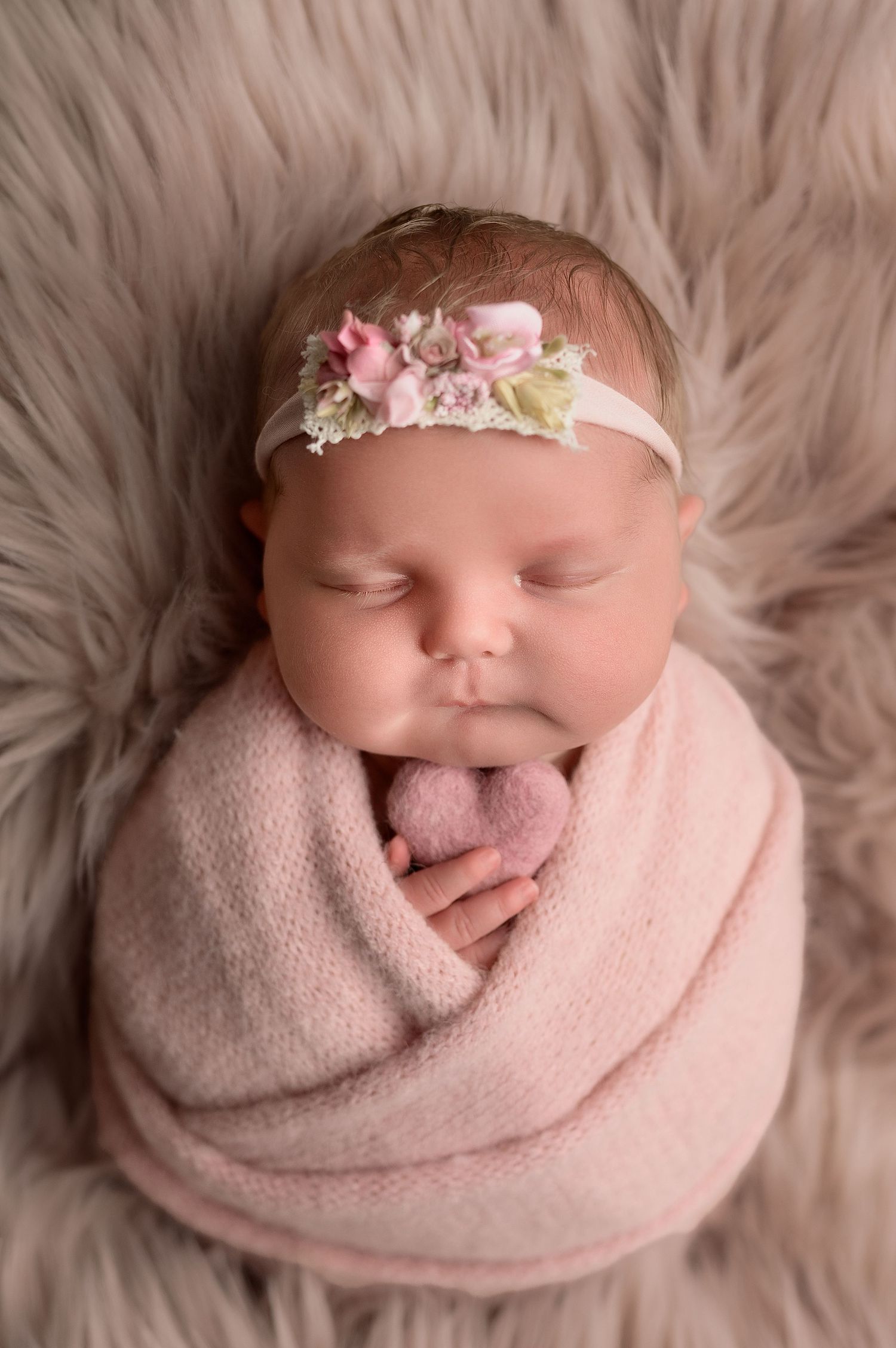 Newborn baby girl wrapped in pink knit blanket with floral headband on fluffy fur backdrop.