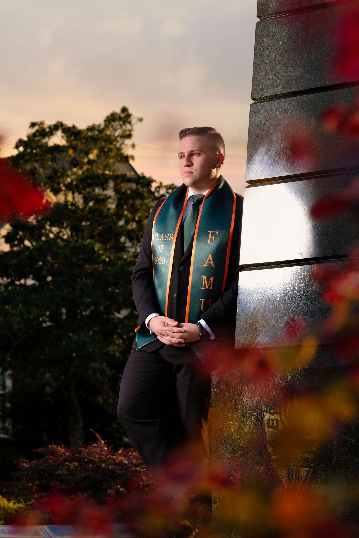 Graduate wearing green and orange stoles over formal attire poses at sunset with autumn foliage in foreground.