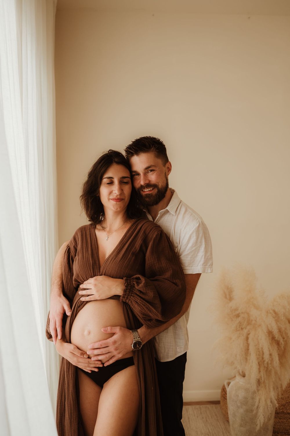 Intimate maternity photo showing a couple embracing near a window with pampas grass decor in a minimalist setting.