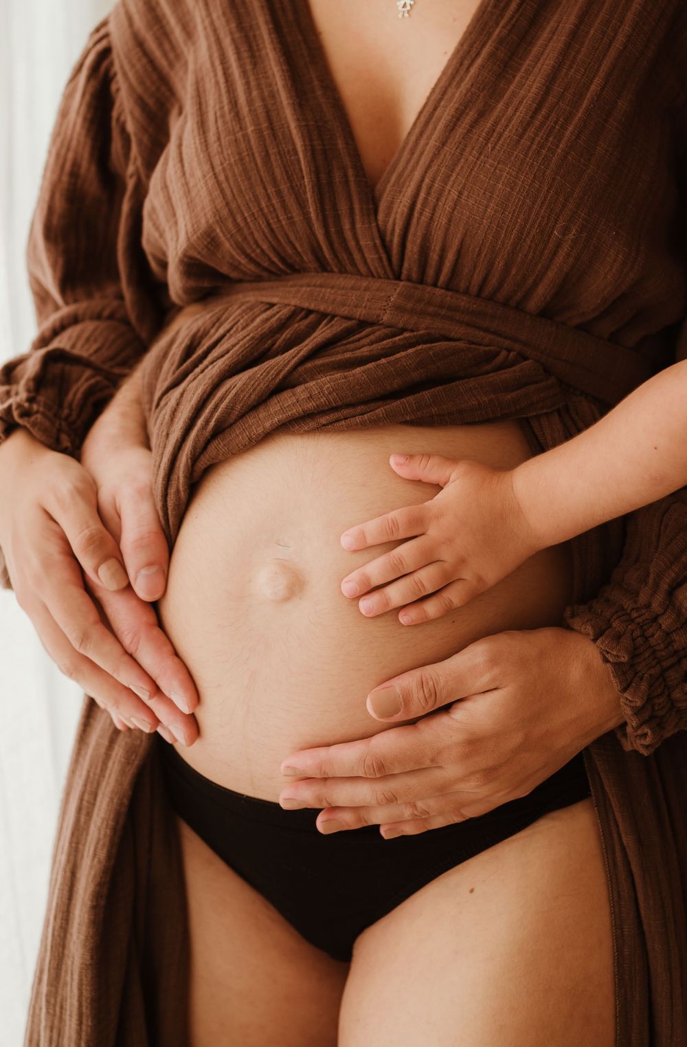 Close up photo of pregnant belly being cradled by hands wearing a brown flowy dress and black underwear.