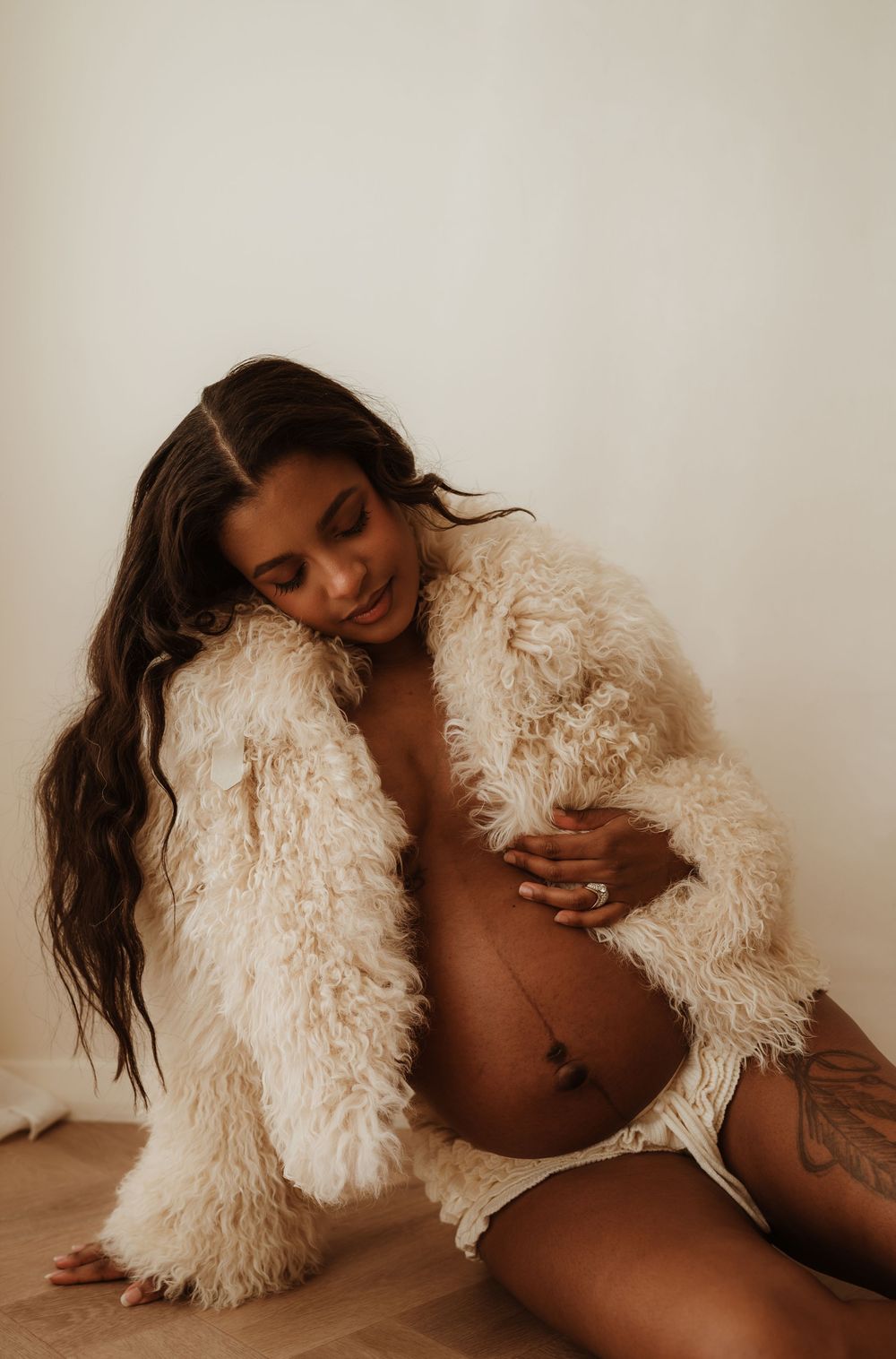 A model in a fluffy cream-colored faux fur jacket poses on wooden flooring in an artistic boudoir photo.
