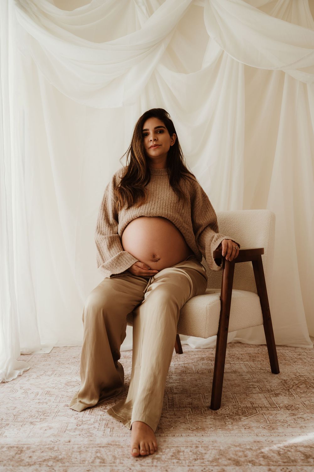 A series of minimalist maternity photos showing a woman in beige loungewear seated on a dark wooden chair against white drapes.